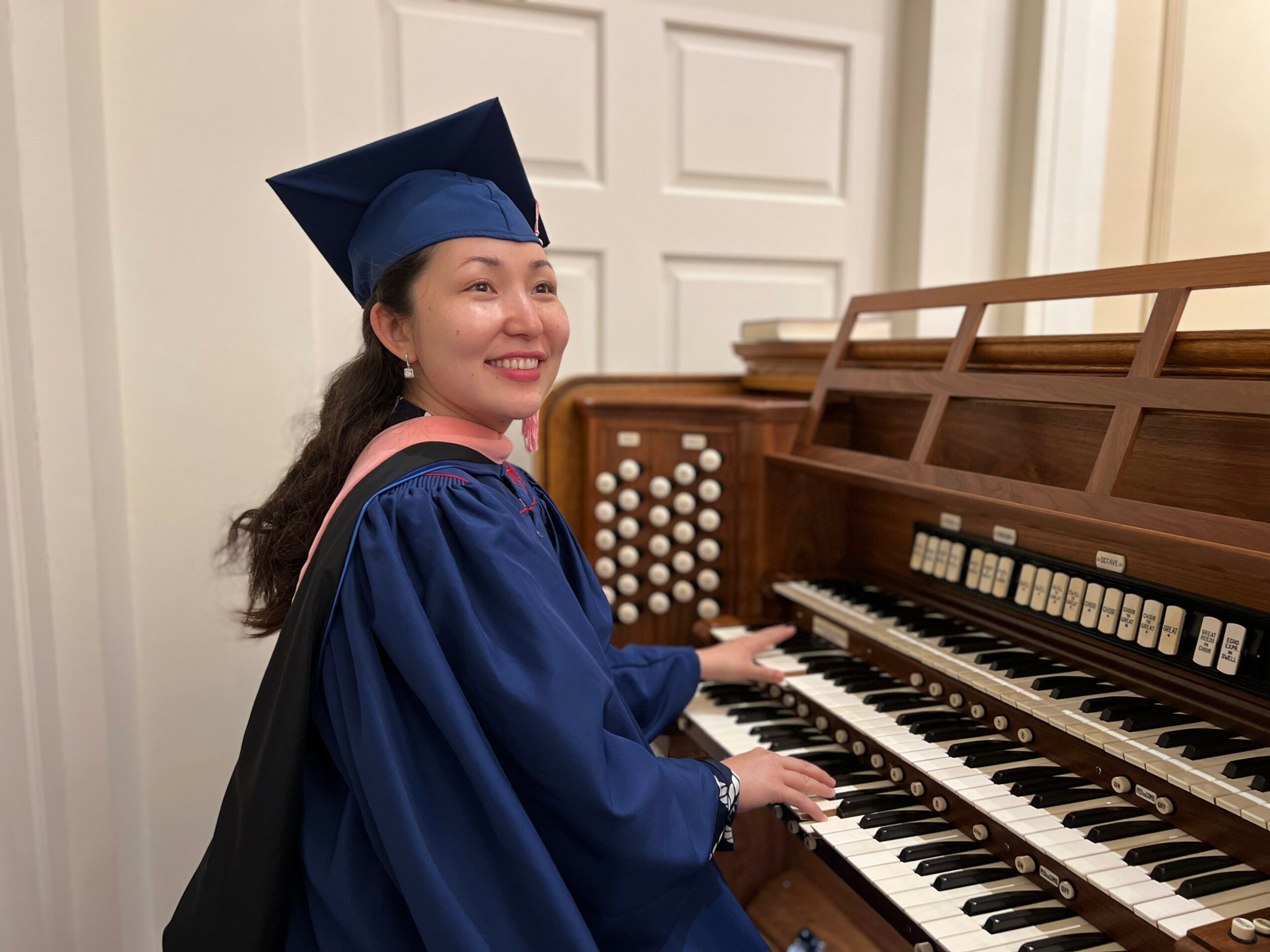 Young woman in graduation gown and cap playing a piano, smiling, in a room with white walls and door.