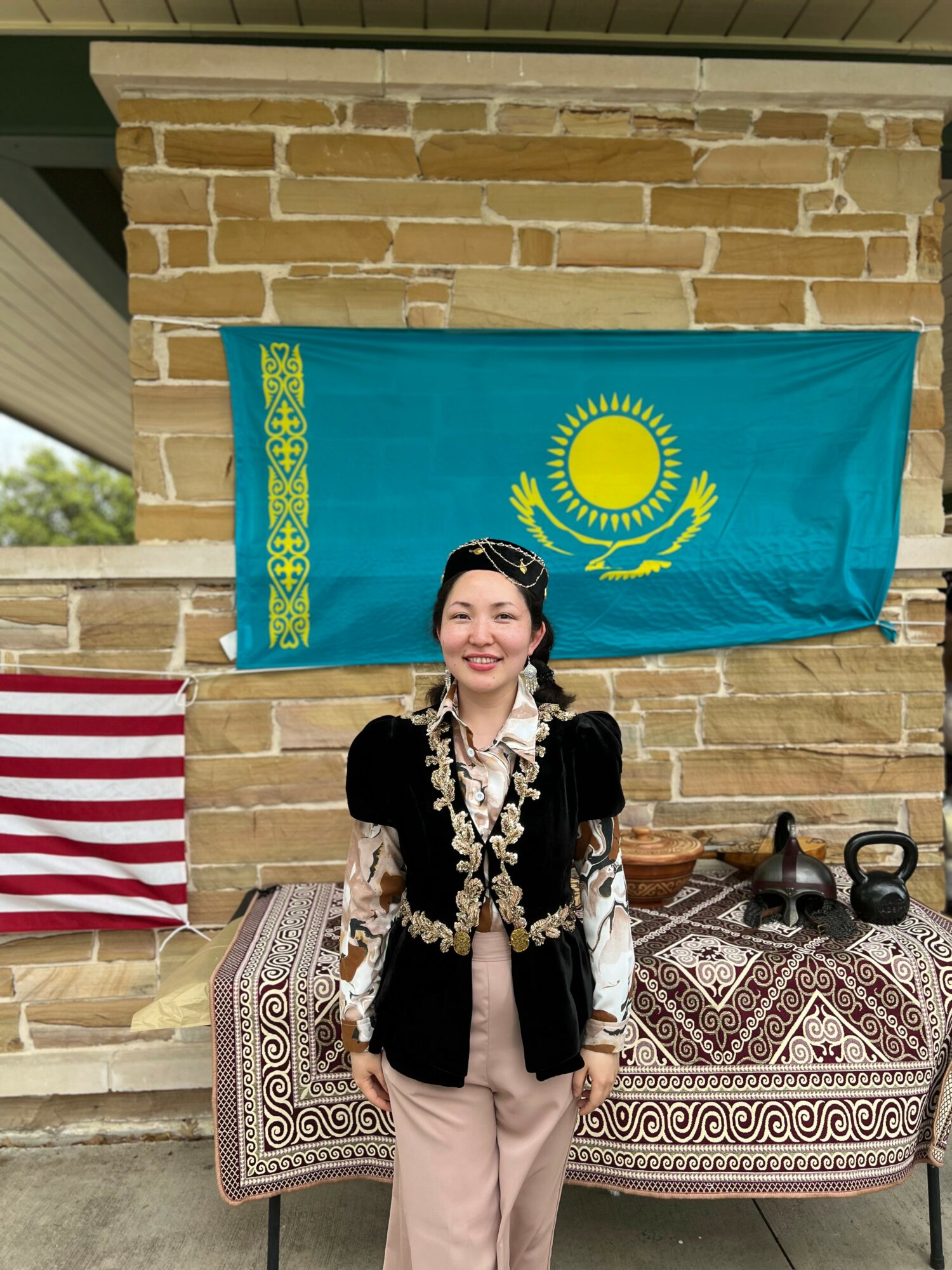 Woman standing in front of a Kazakh flag and a striped cloth, smiling, with a table and kettlebells nearby.