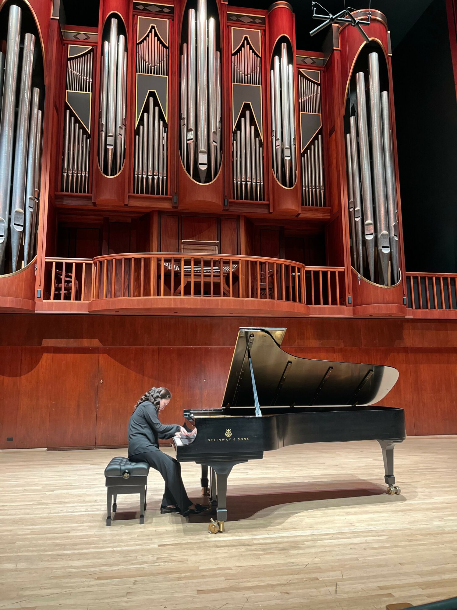 Person playing grand piano on stage with large pipe organ in background.