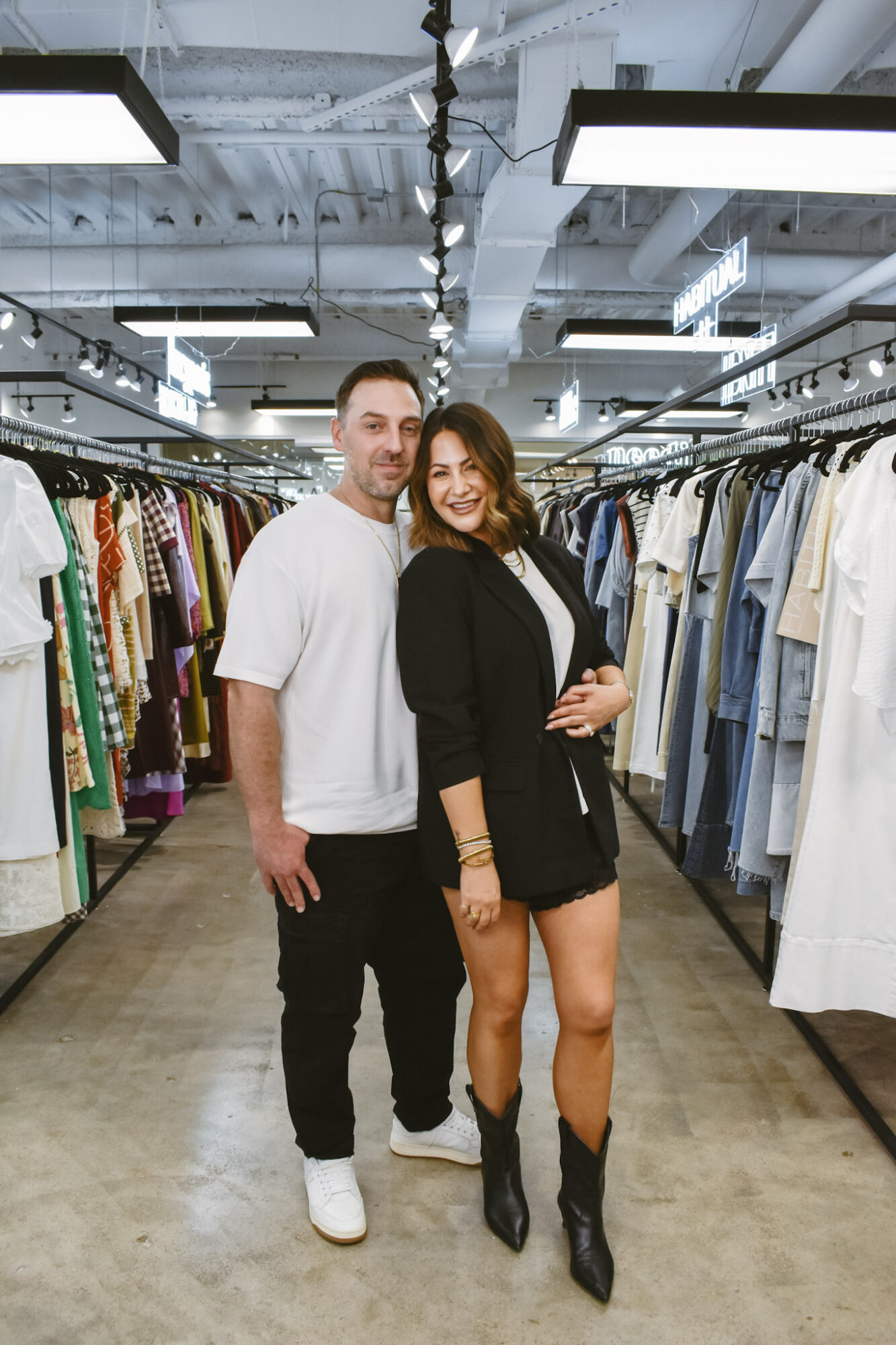 A man and woman stand together in a clothing store aisle, smiling, surrounded by racks of clothes.
