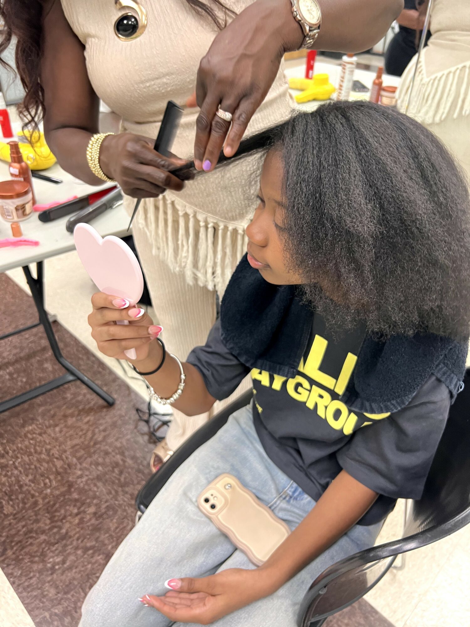 Young woman with curly hair holding a pink mirror, sitting at a table with a phone on her lap.