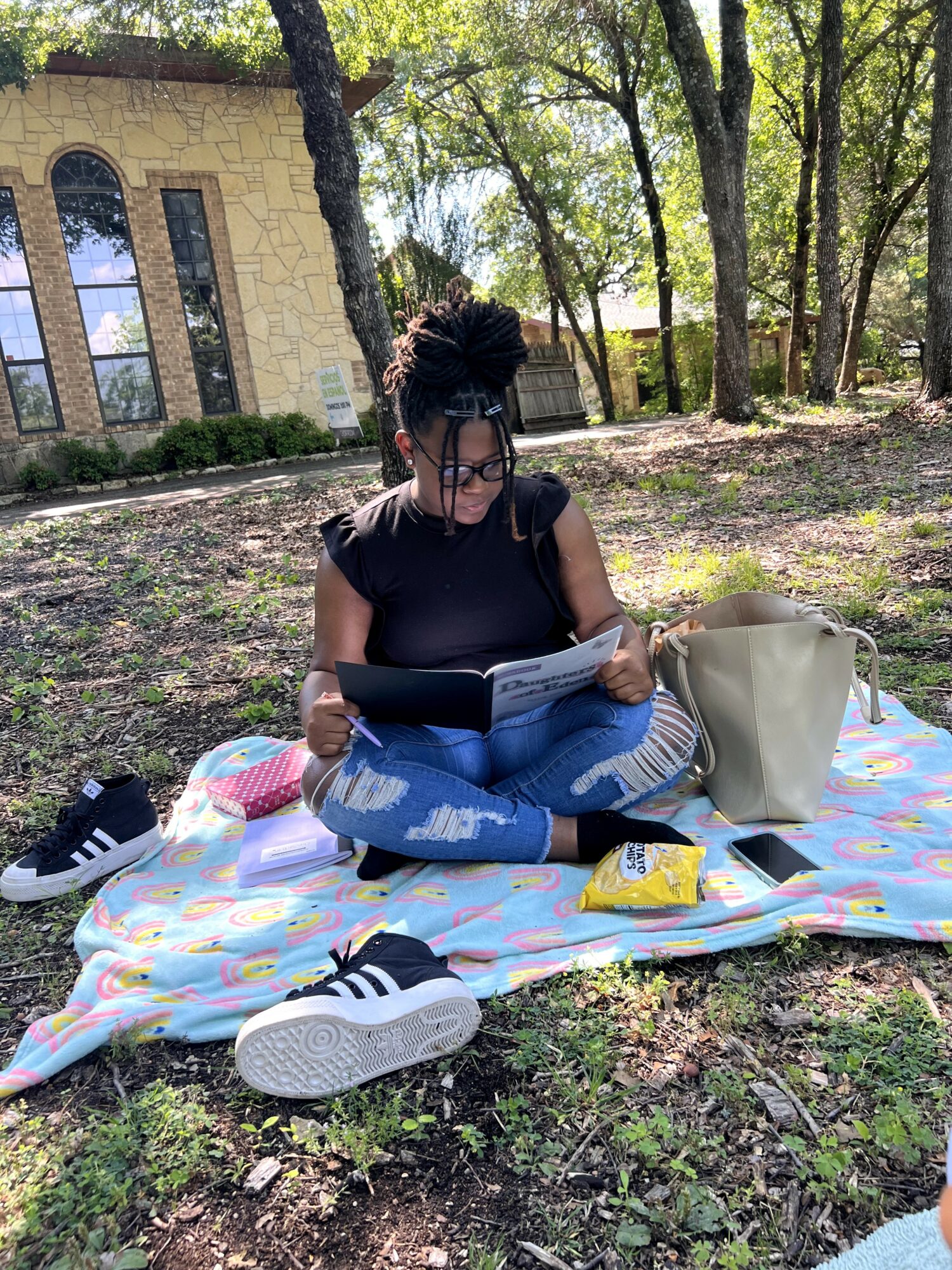 Person sitting on a colorful blanket reading a book outdoors with trees and a building in background.