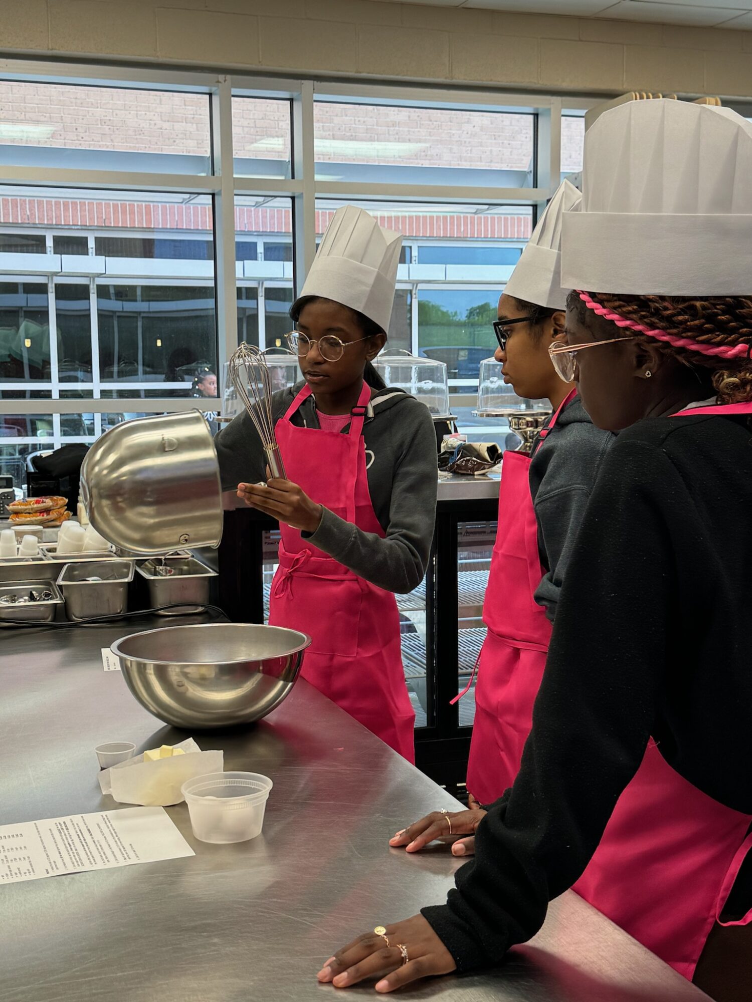 Three children wearing chef hats and aprons in a kitchen, one pouring from a pot into a bowl, others watching.