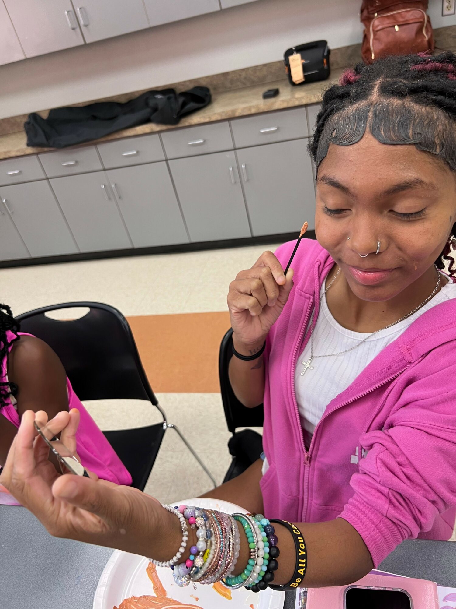 Girl with braided hair and colorful bracelets on her arm, smiling and looking at her hand, in a classroom setting.