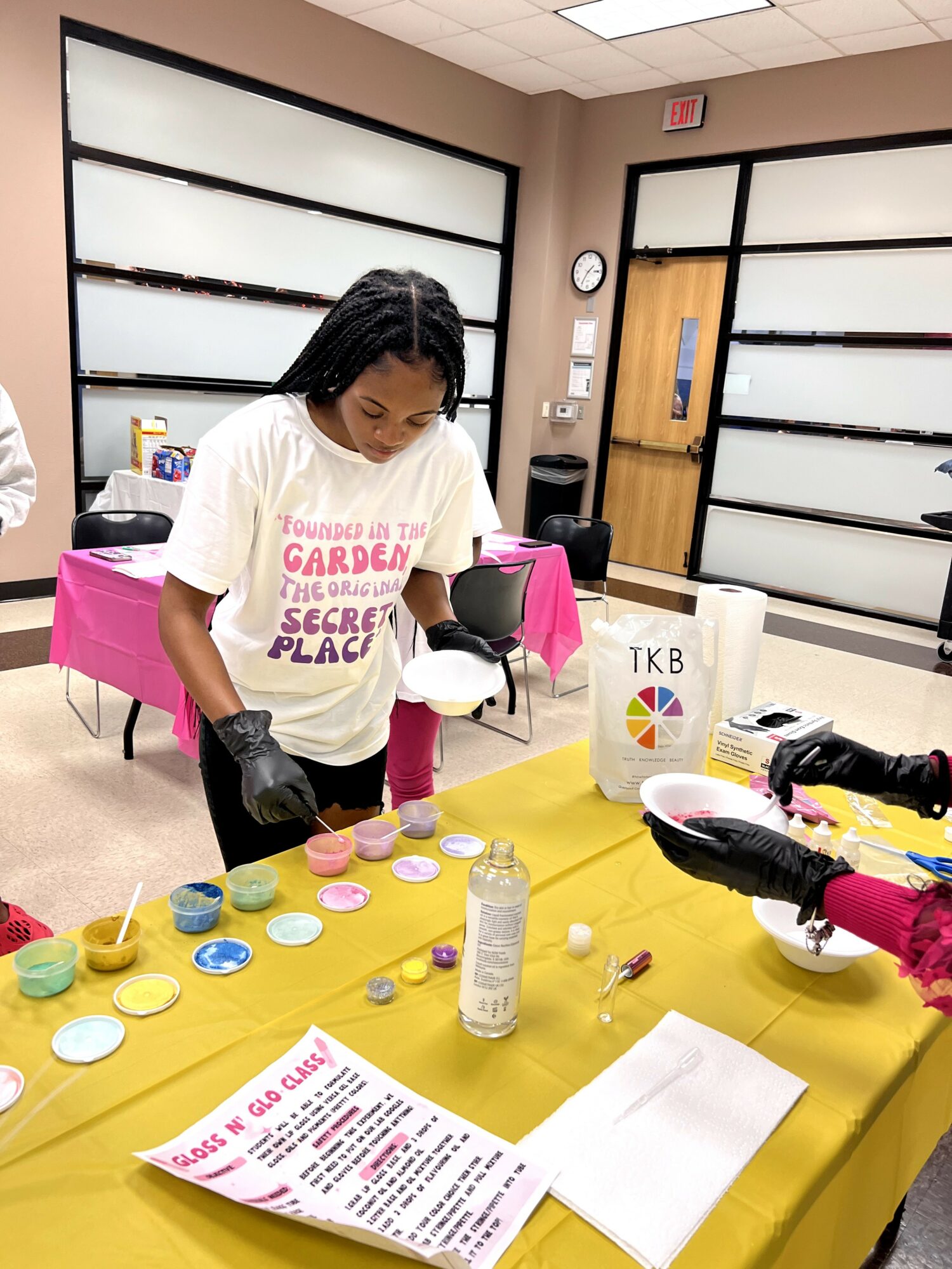 Woman wearing gloves decorating cupcakes at a table with colorful frosting and supplies in a room.