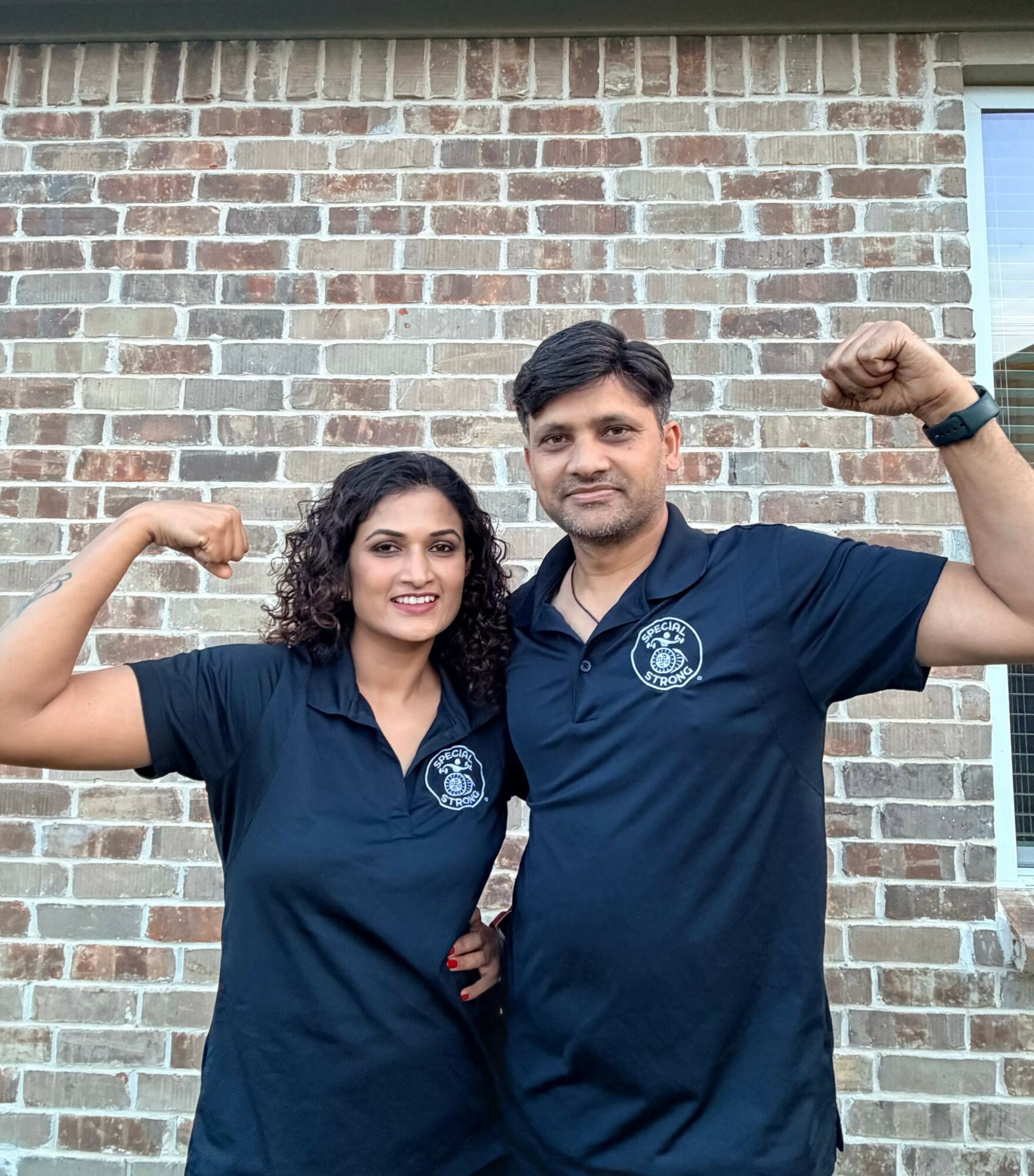 Two people in navy blue polo shirts flexing their biceps outdoors against a brick wall.