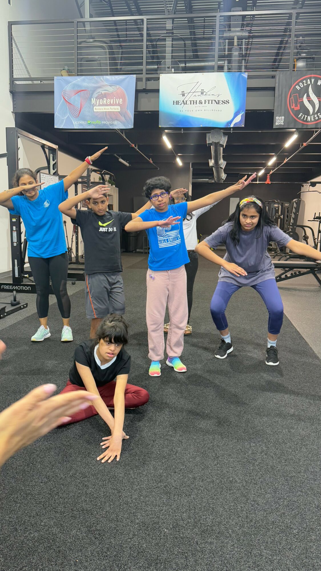 Five people in gym, four standing and one sitting on the floor, posing with arms outstretched or on hips.