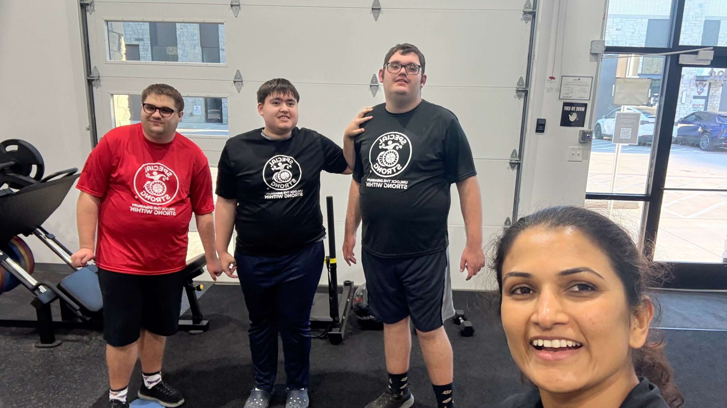 Four people in a gym, three standing and one taking a selfie, all smiling, with gym equipment in background.