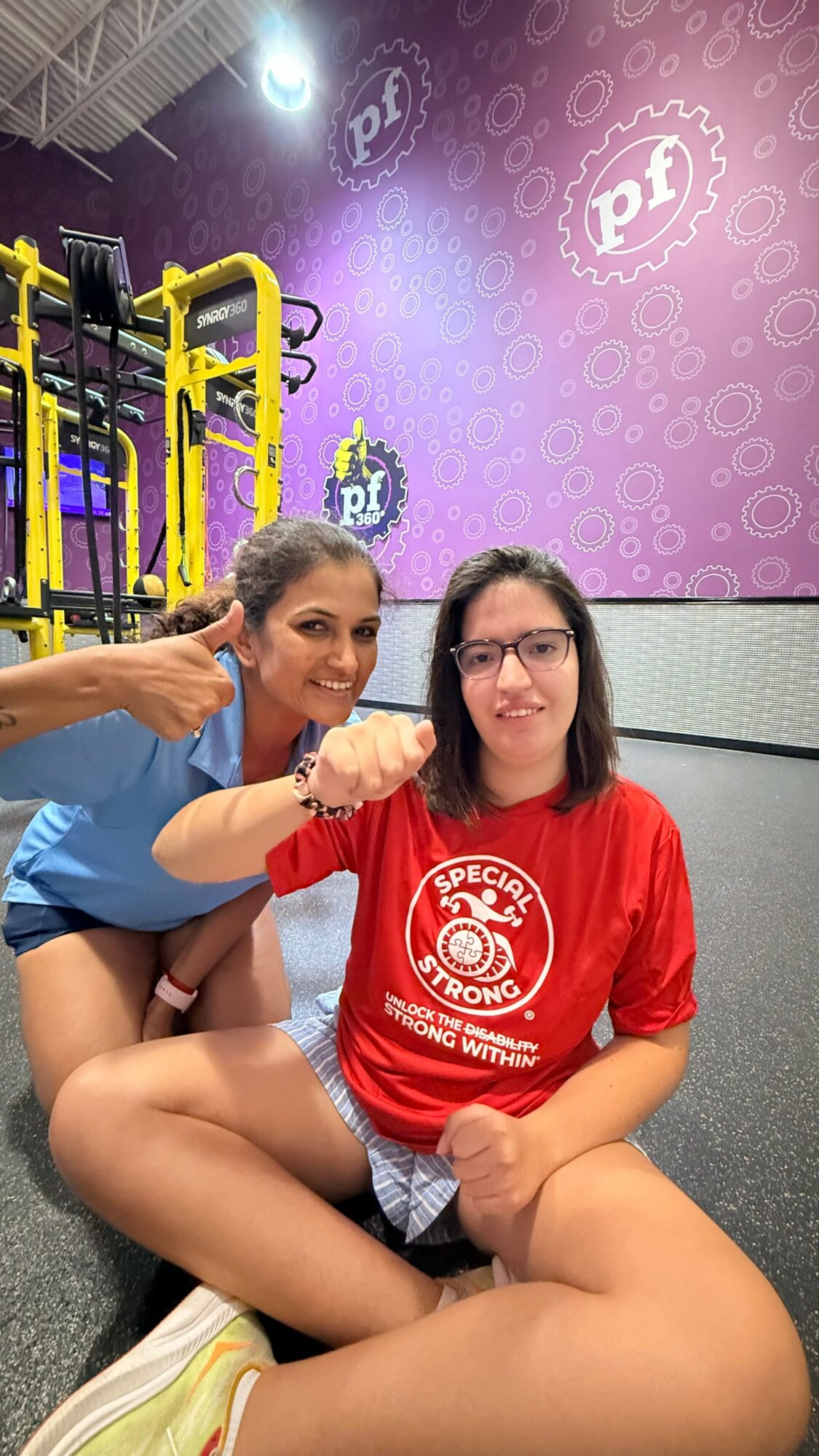 Two women sitting together, one giving a thumbs-up, smiling, in a colorful indoor setting with purple walls and yellow structures.