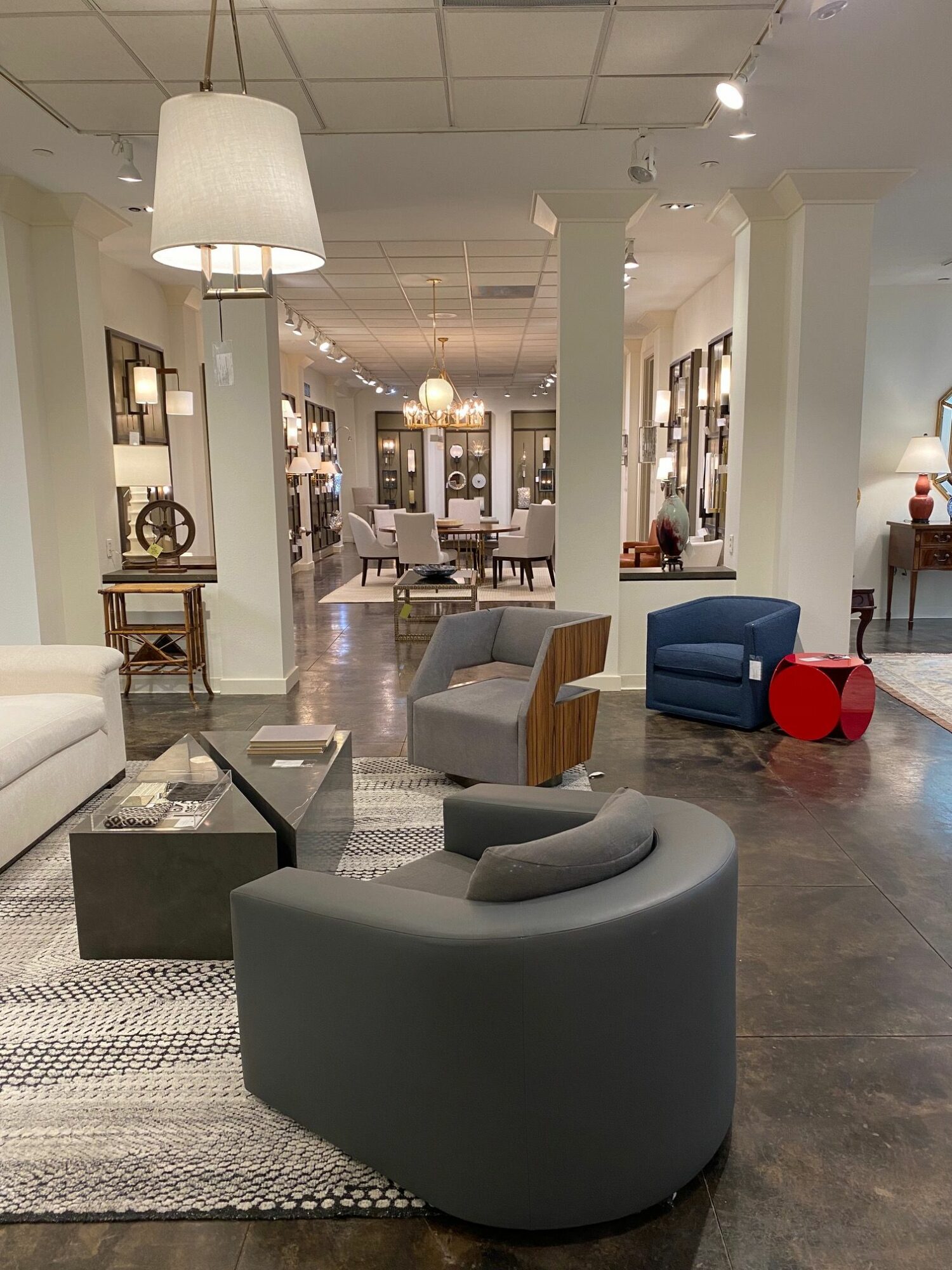 Modern furniture in a store, including a gray rounded chair, a gray armchair, and a blue armchair with a red side table.
