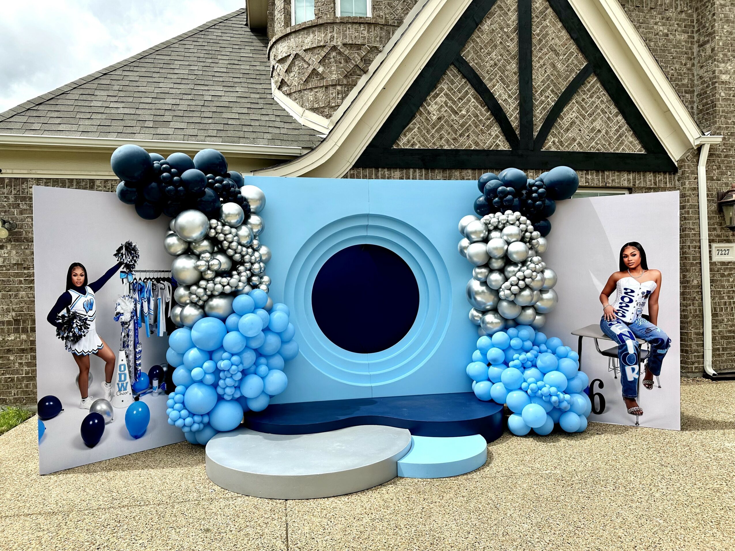 Decorative backdrop with blue, black, and silver balloons, circular stage, and two large portrait displays on sides.