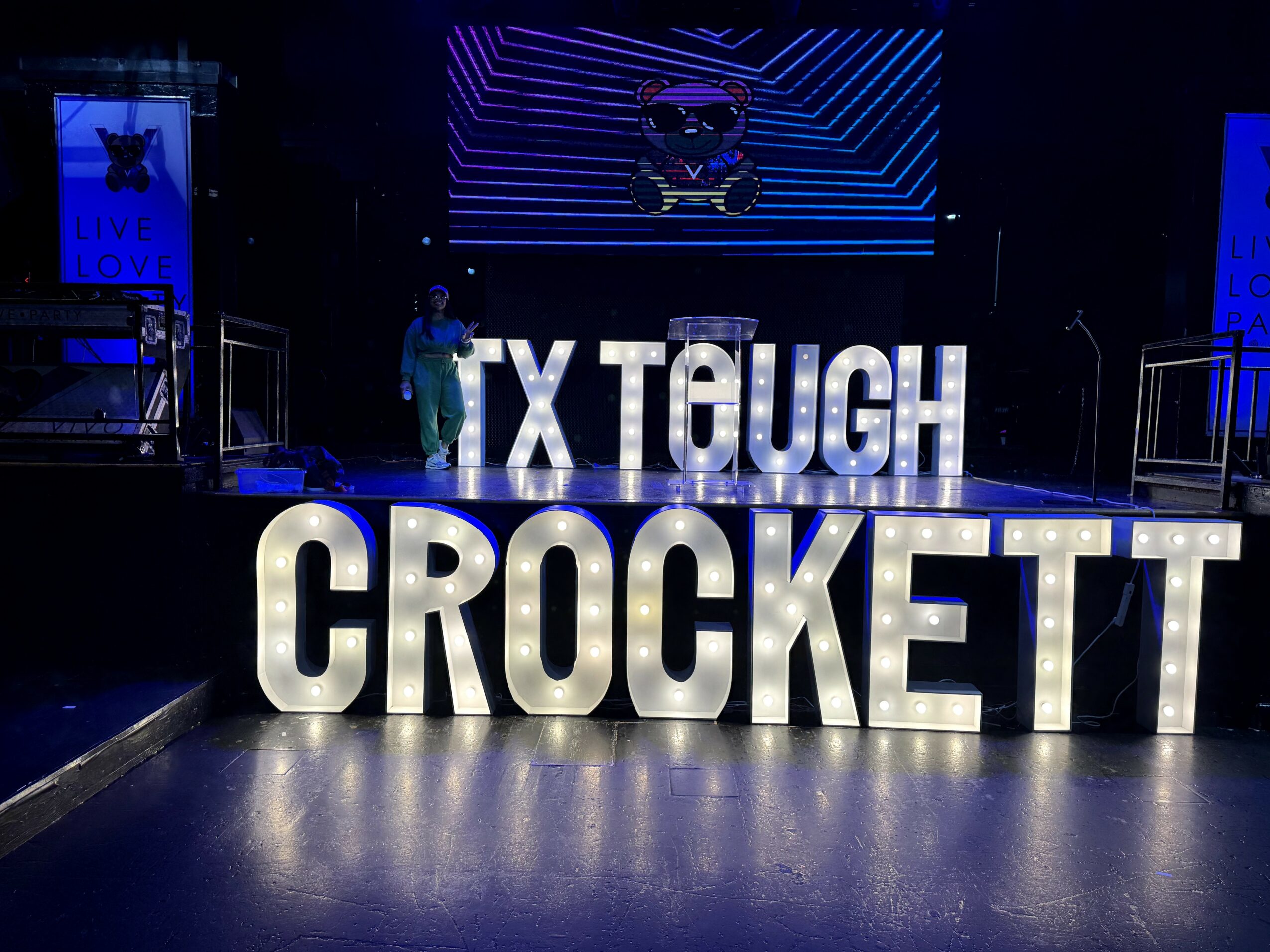 Stage with large illuminated letters spelling 'CROCKETT' and partially visible text 'TX TEUGH'. Person standing on stage.