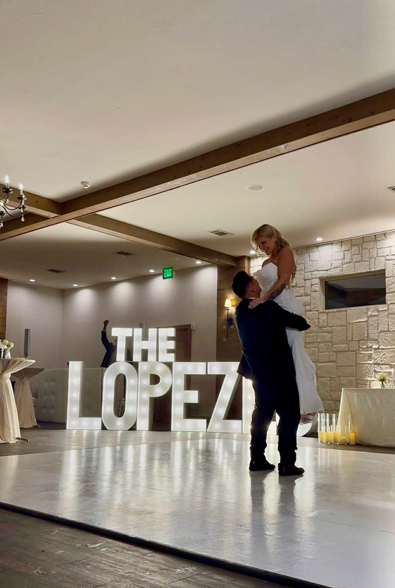 Person lifting a child in a room with large illuminated letters spelling 'THE LOPEZ'.