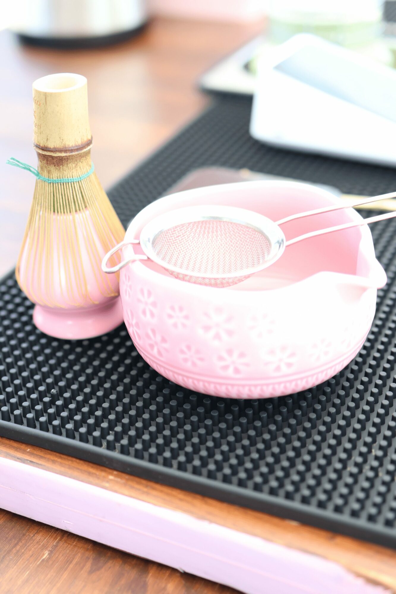 Pink bowl with a strainer on top, next to a small decorative bottle, on a black textured surface.