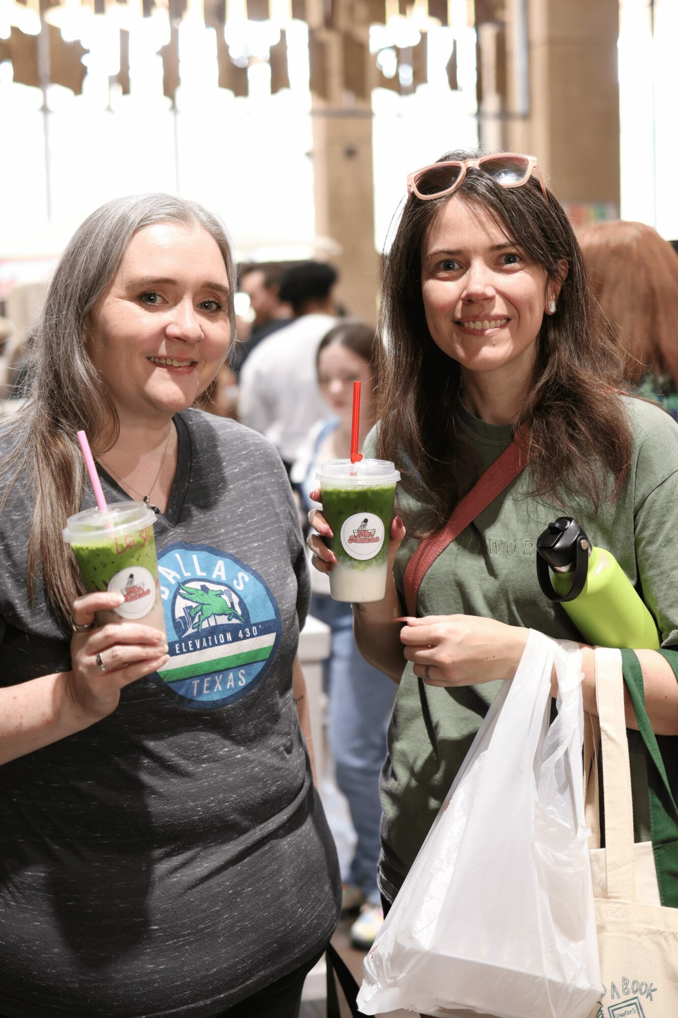 Two women smiling and holding drinks with straws in a crowded indoor setting.
