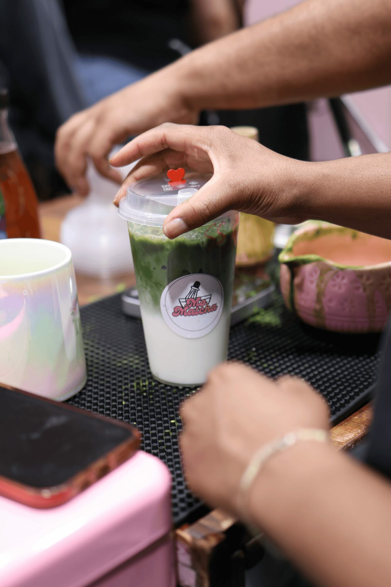 Person holding a clear cup with layered green and white drink, lid with straw, on a black surface.