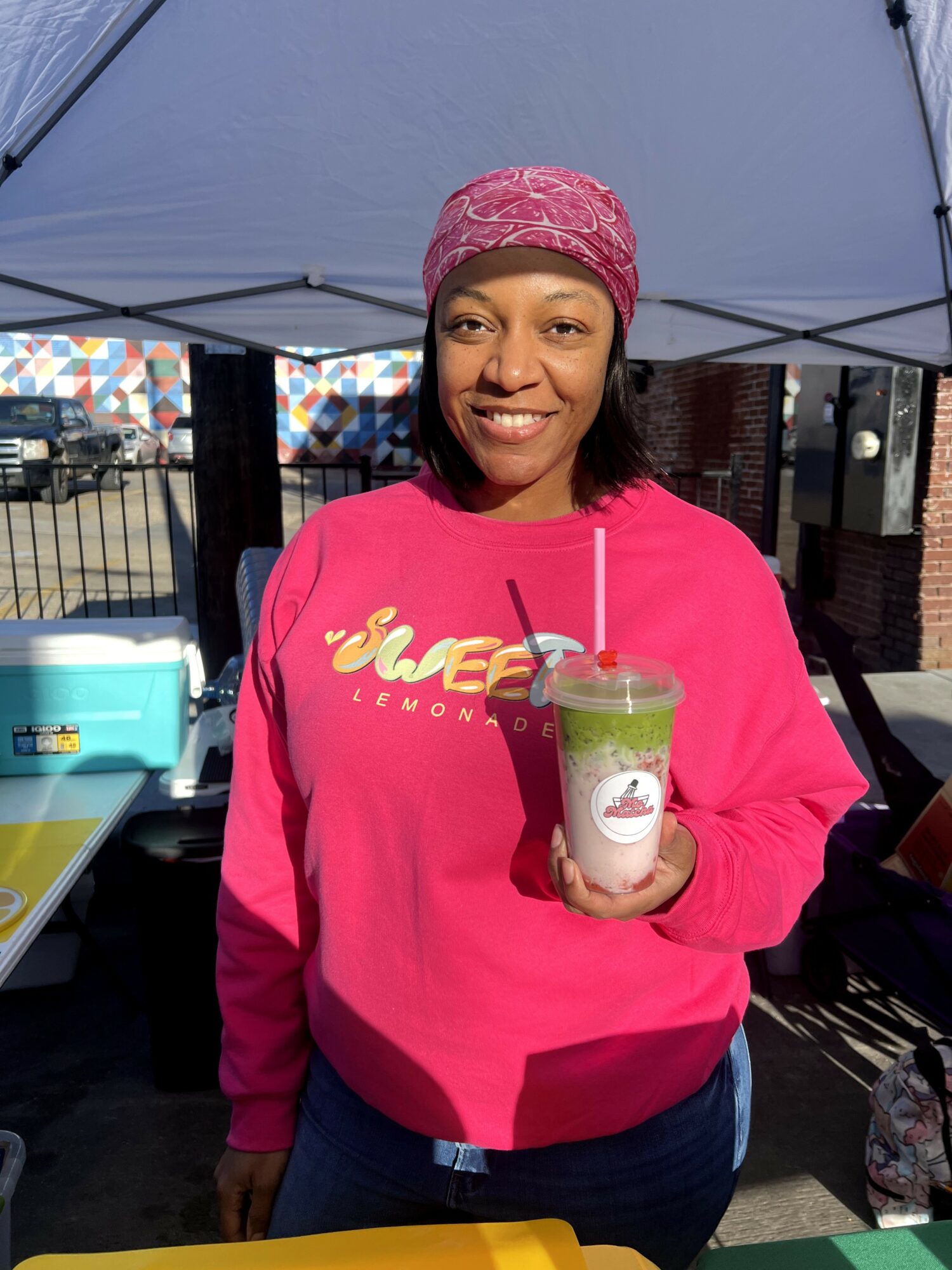 Woman in pink sweatshirt and pink headscarf holding a drink with a straw, smiling outdoors under a canopy.