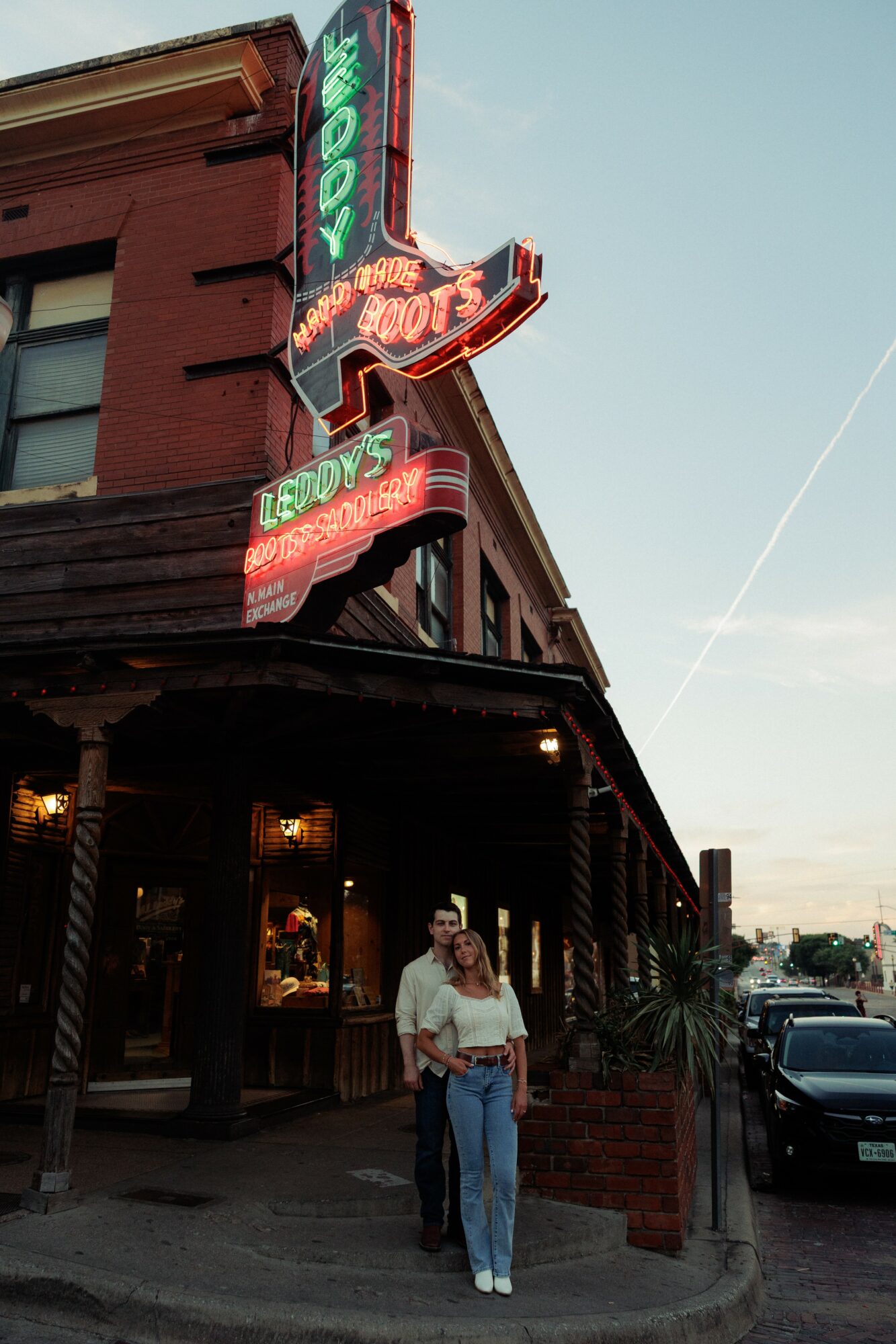 Two people stand outside a building with neon signs, under a covered porch, during daytime.