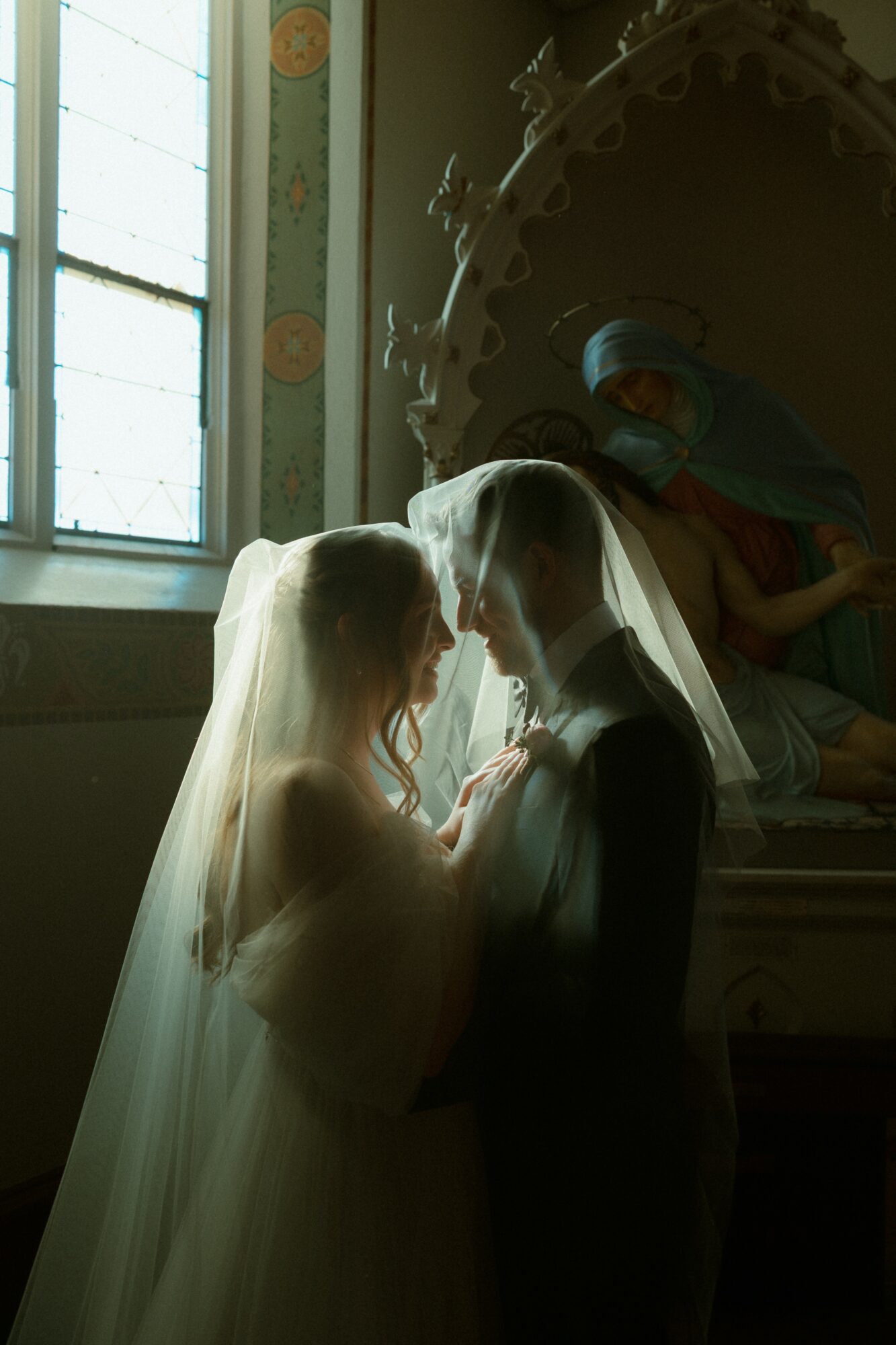 Two women in wedding dresses and veils face each other inside a church, with a statue and stained glass window behind them.