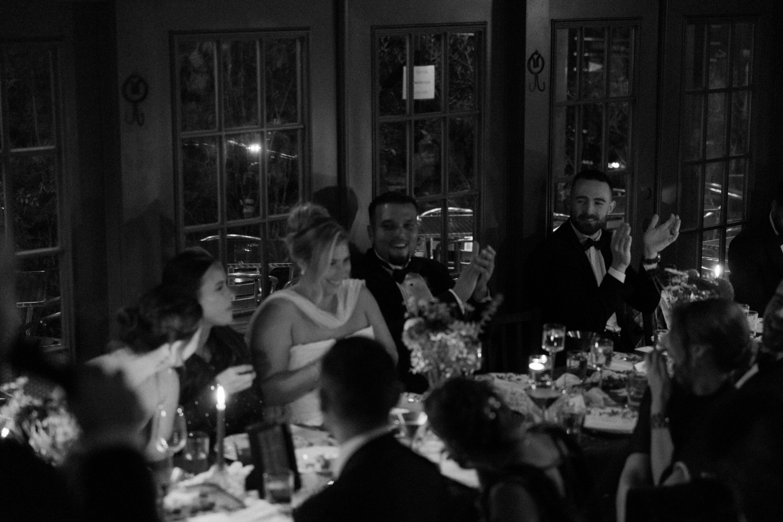 People sitting around a table at a dinner event, some clapping and talking, in a room with large windows.