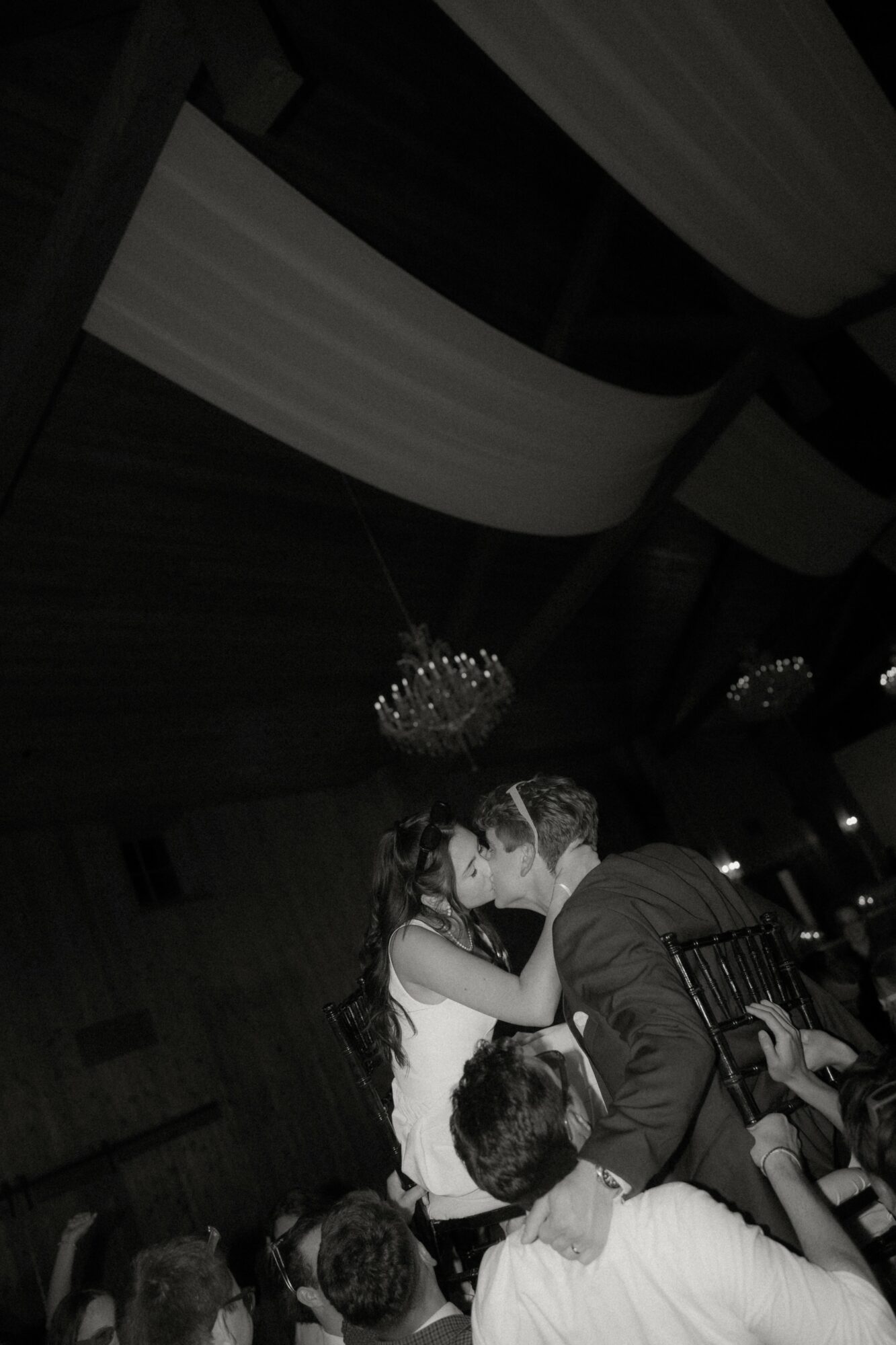 Two people kiss on a dance floor with a chandelier and draped fabric ceiling in the background.