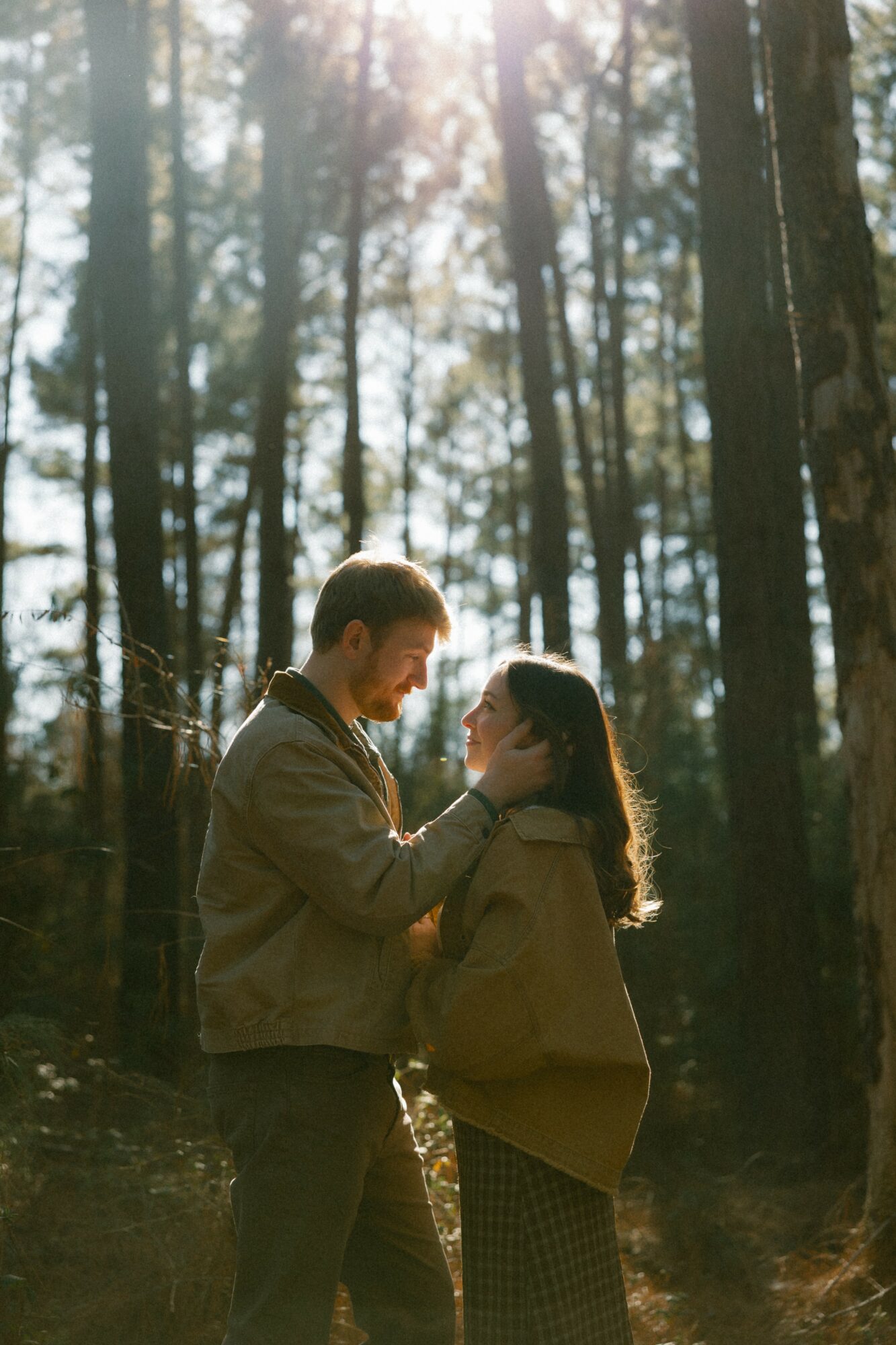 A couple stands close in a forest, gazing at each other with sunlight filtering through trees behind them.