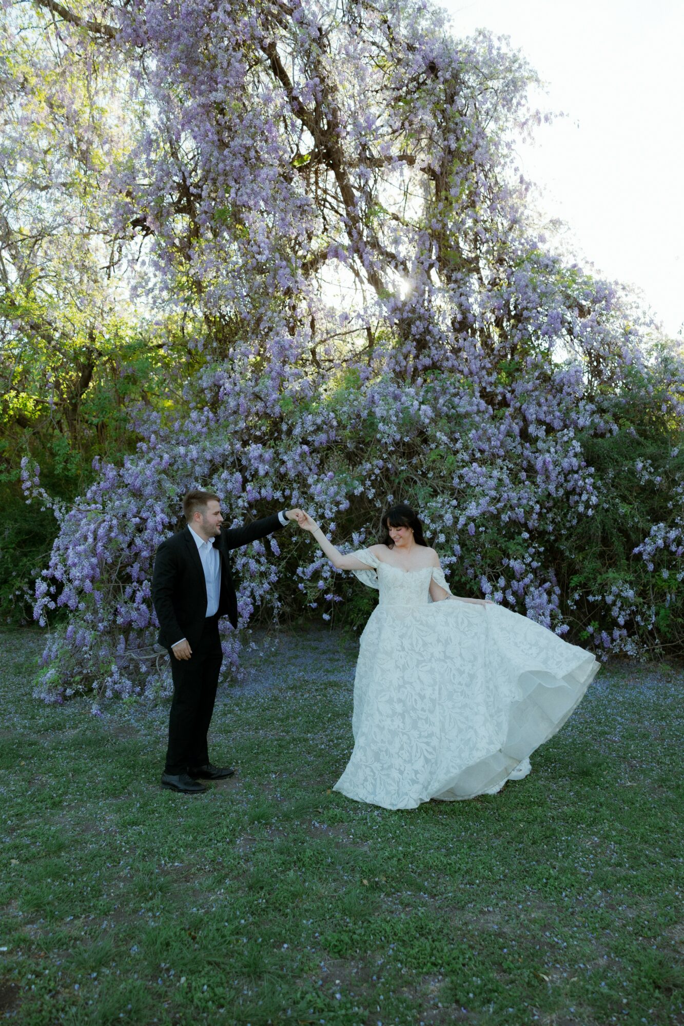 Couple in wedding attire under a large flowering tree, holding hands outdoors.