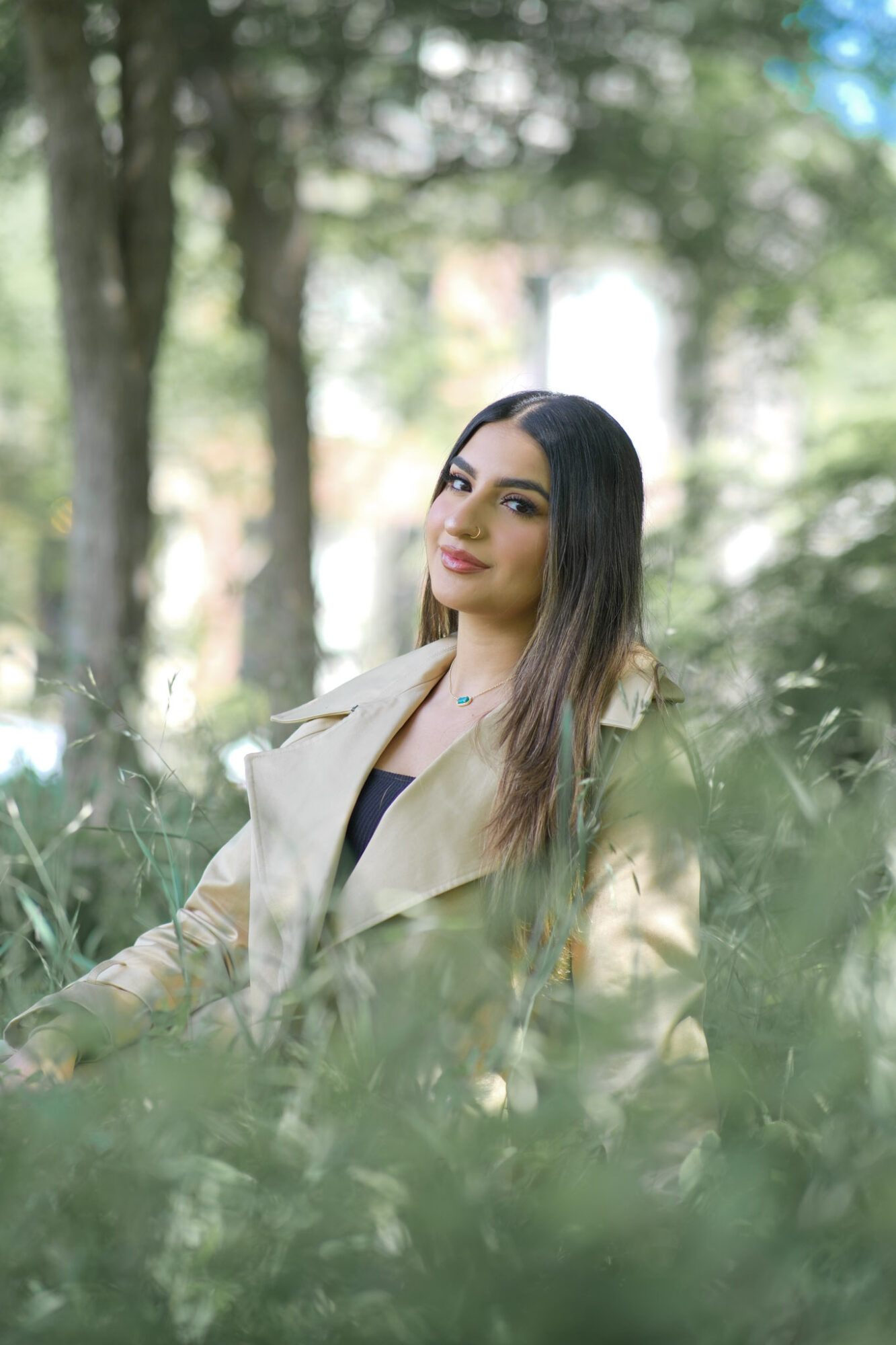Young woman with long dark hair and makeup, wearing a beige coat outdoors among trees and greenery.
