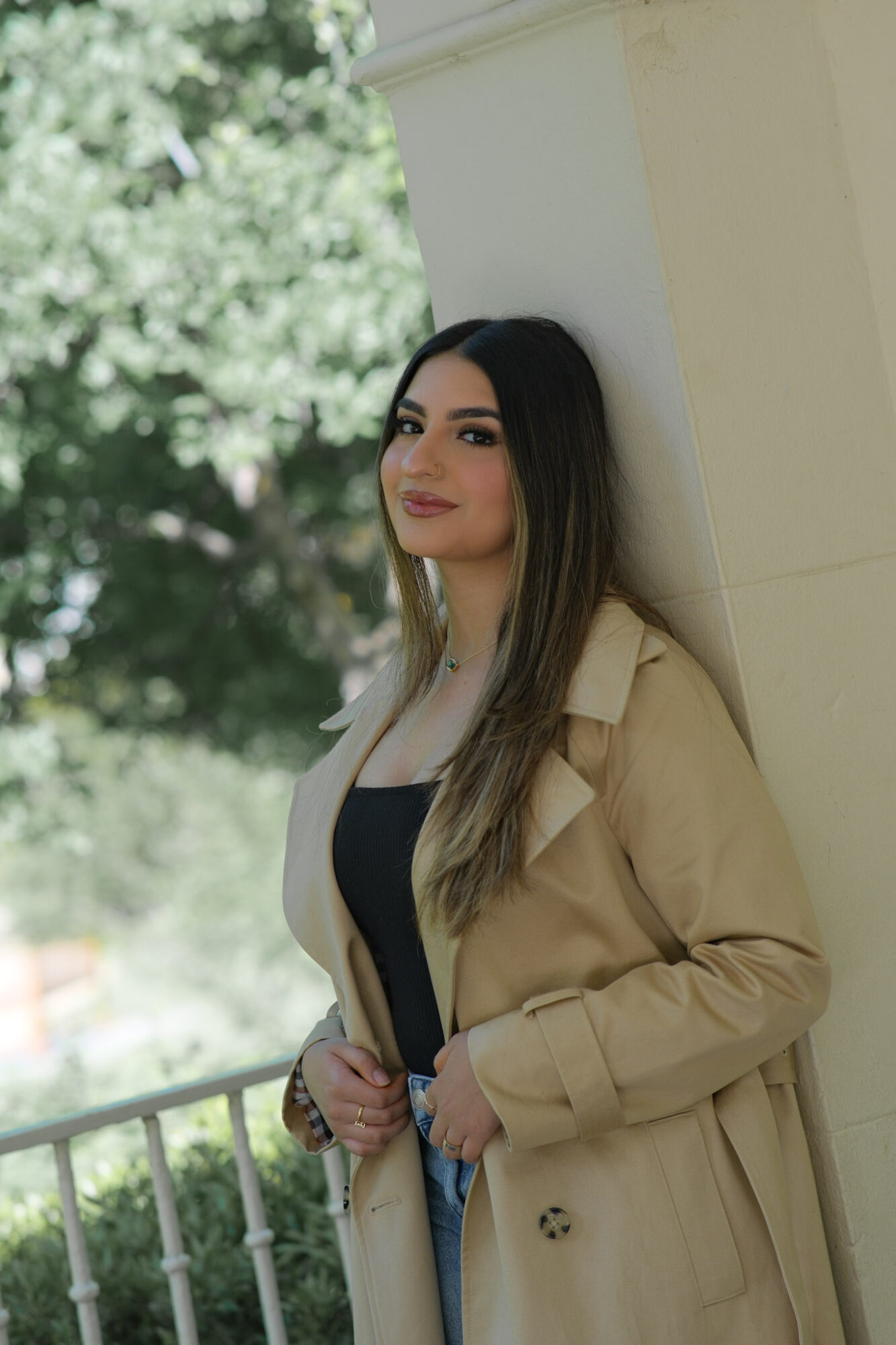 Young woman with long dark hair leaning against a wall outdoors, smiling, wearing a beige coat and black top.
