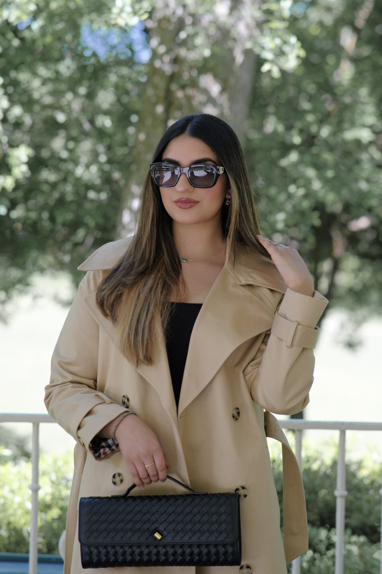 Woman with long hair wearing sunglasses and a beige trench coat outdoors, holding a black handbag.