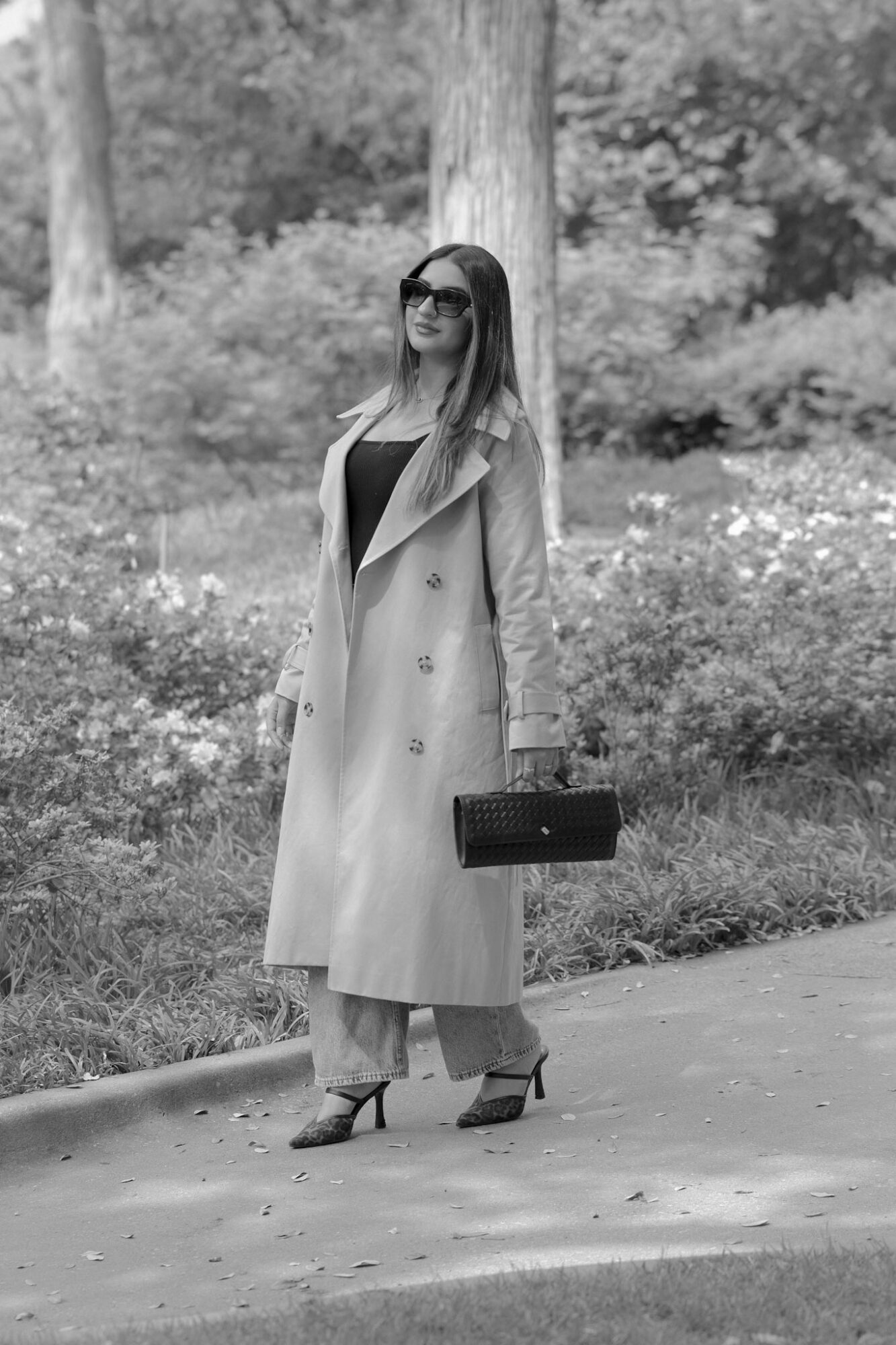 Woman standing outdoors in a park, wearing sunglasses, a long coat, and holding a handbag.
