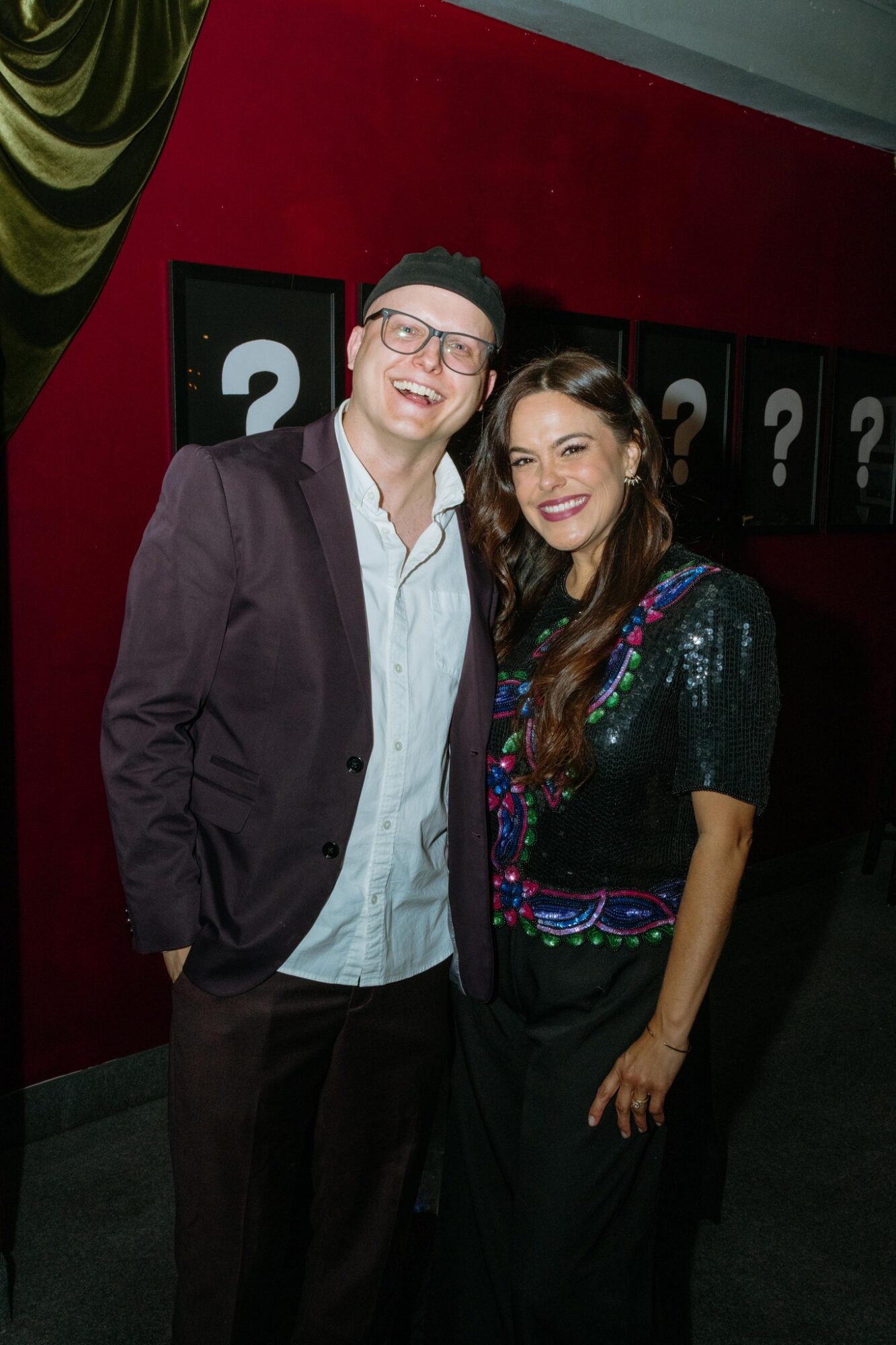Two people smiling, standing close together in front of a red wall with question marks, indoors.