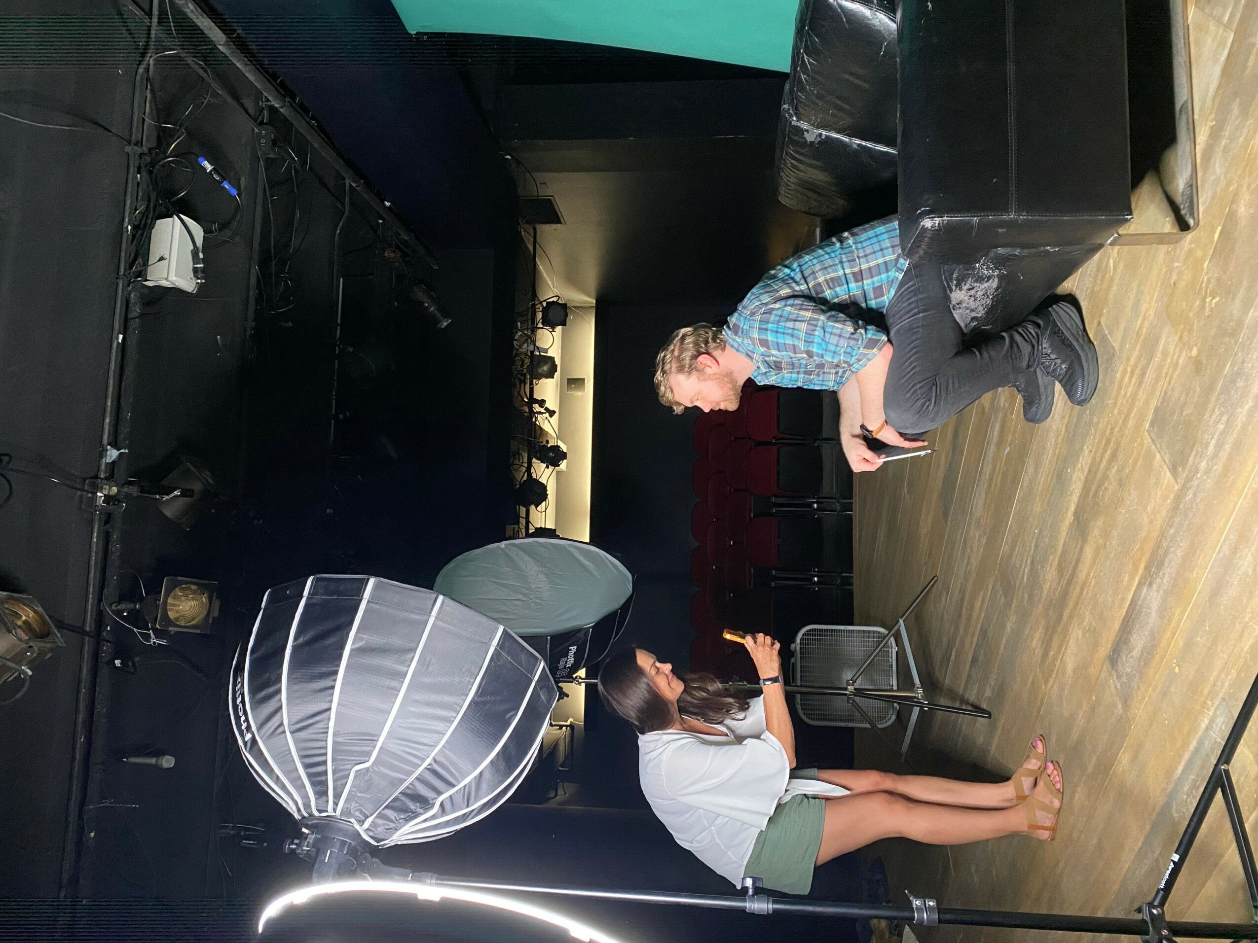 Two people in a room with black walls and wooden floor, one sitting on a chair and the other standing, both using phones.