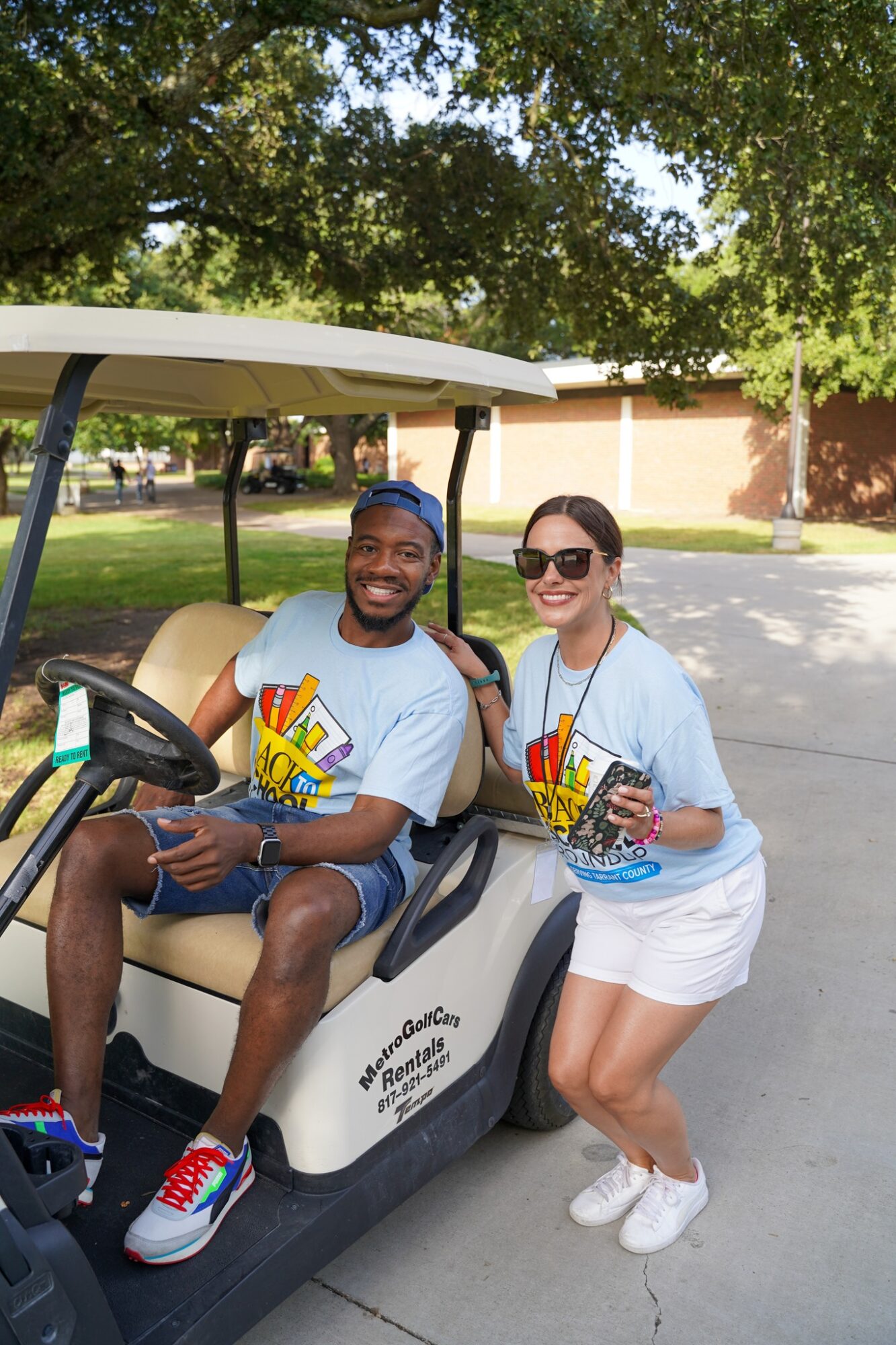 Two people sitting on a golf cart outdoors, smiling, holding drinks, with trees and a building in background.