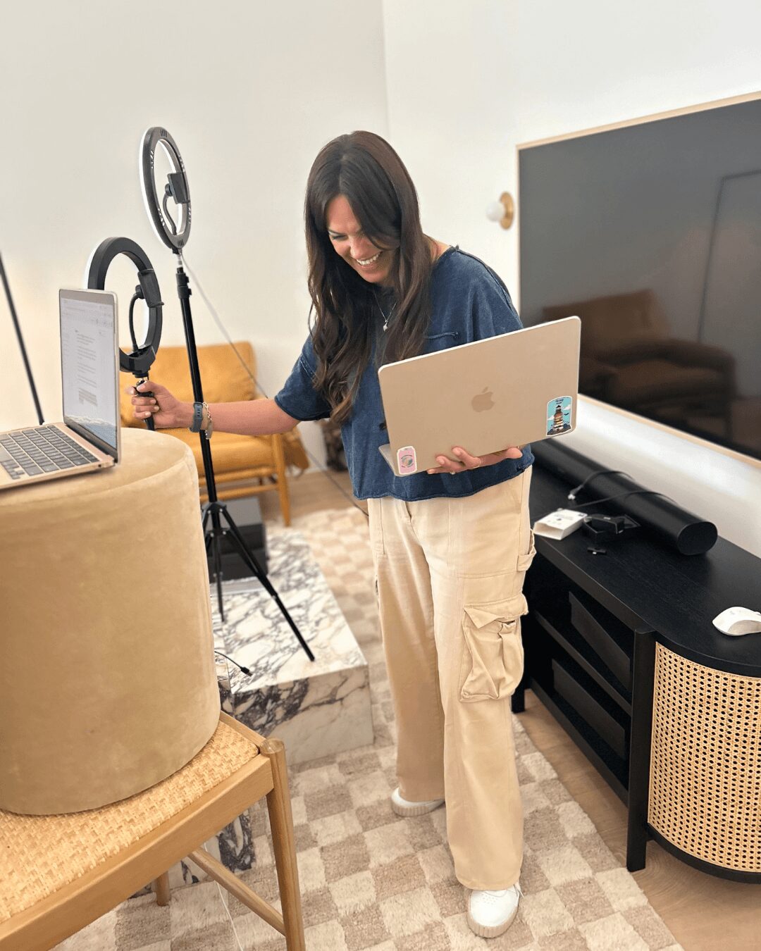 Woman holding a laptop and smiling in a room with a large TV, ring light, and a chair.