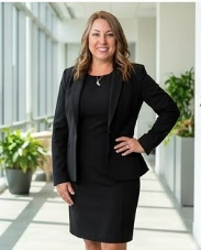 Woman in black suit standing in bright corridor with plants, smiling, hand on hip.