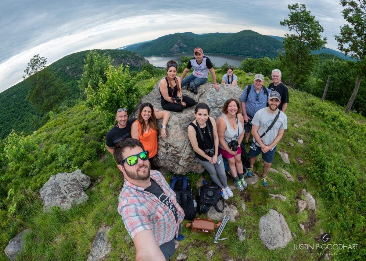Group of people outdoors on grassy hill with trees and mountains in background, taking a selfie.