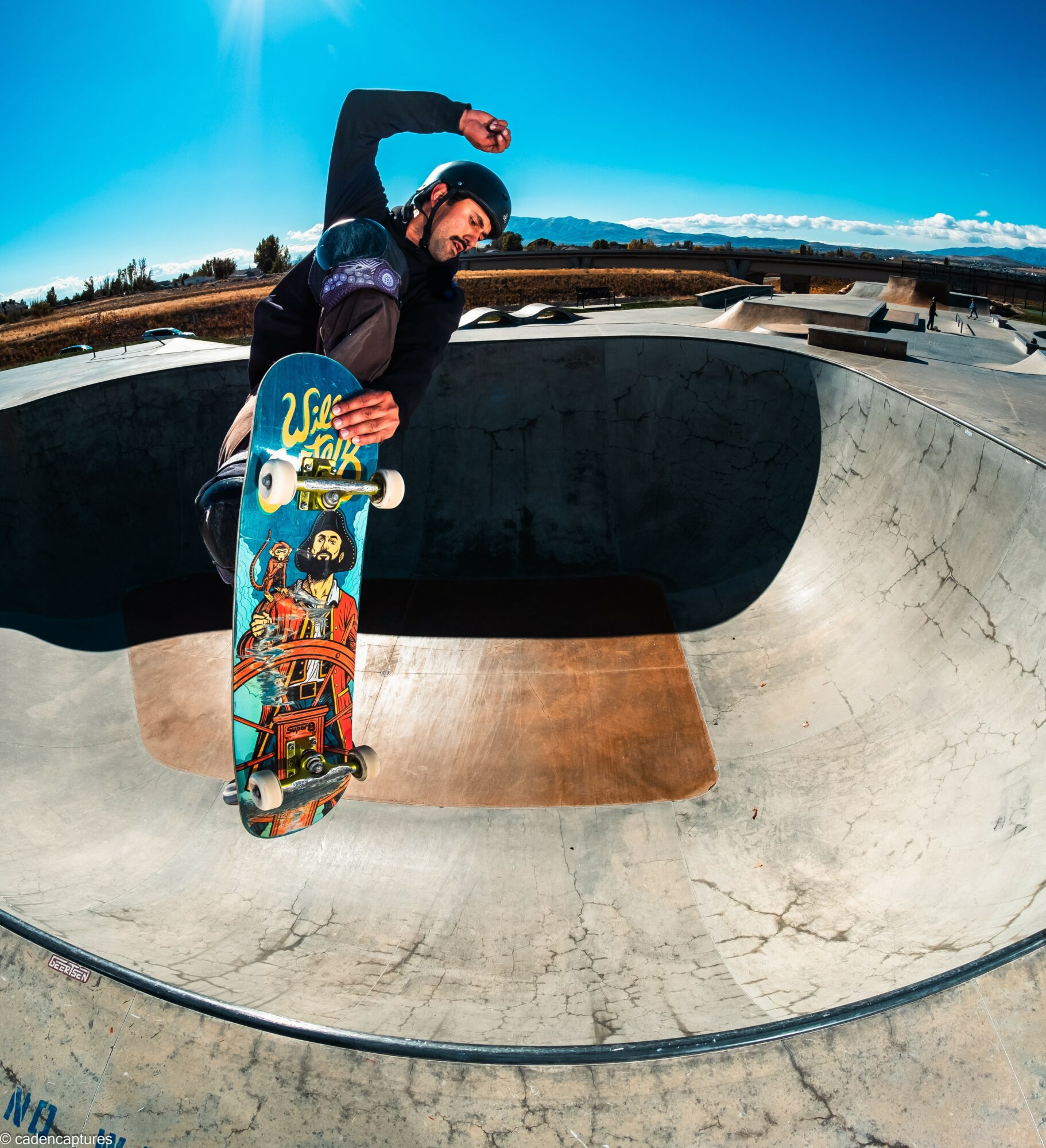 Skateboarder in a skate park bowl holding skateboard, wearing helmet and gloves, with blue sky and mountains in background.
