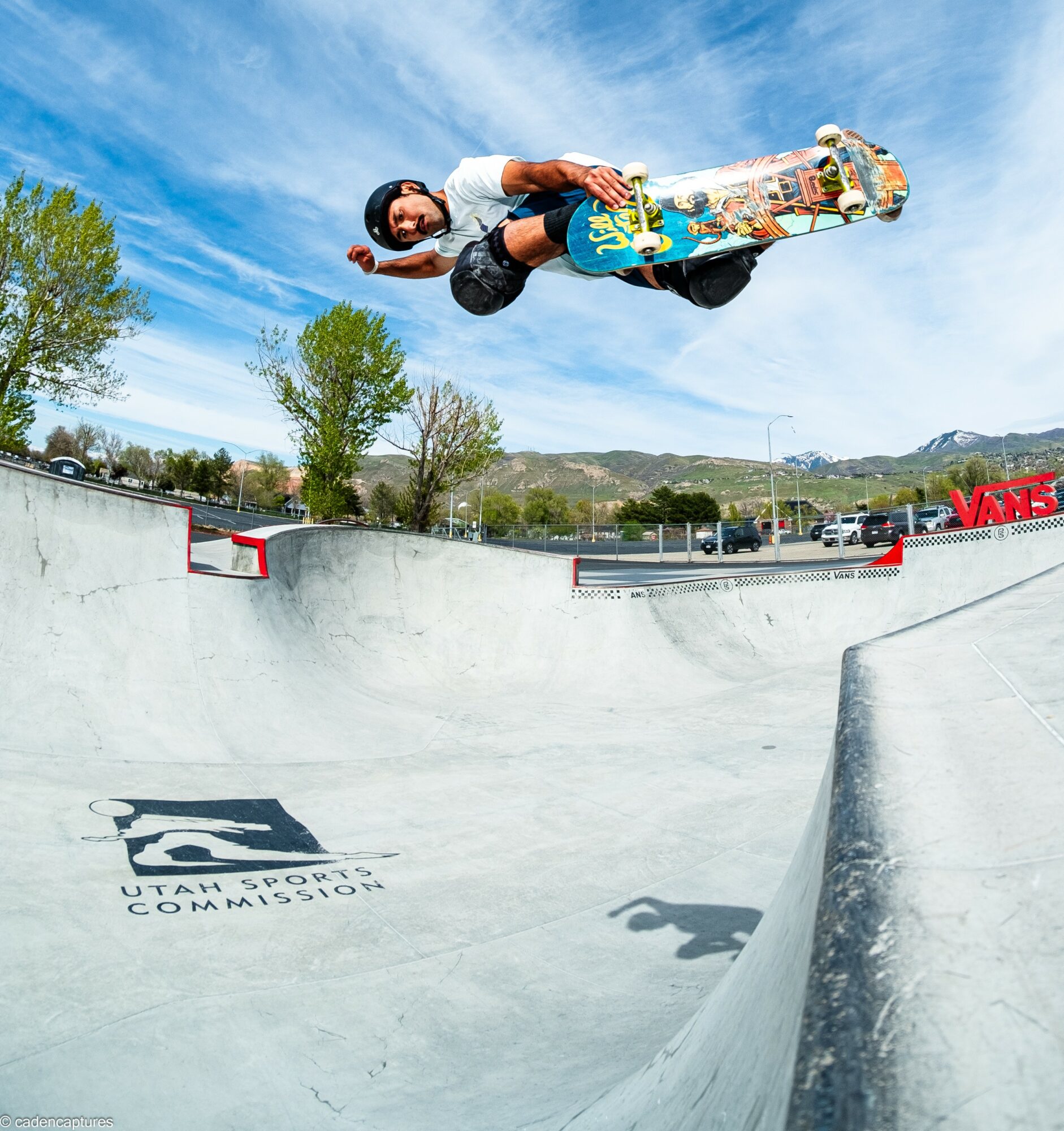 Skateboarder performs a trick in a skatepark with trees and blue sky in the background.
