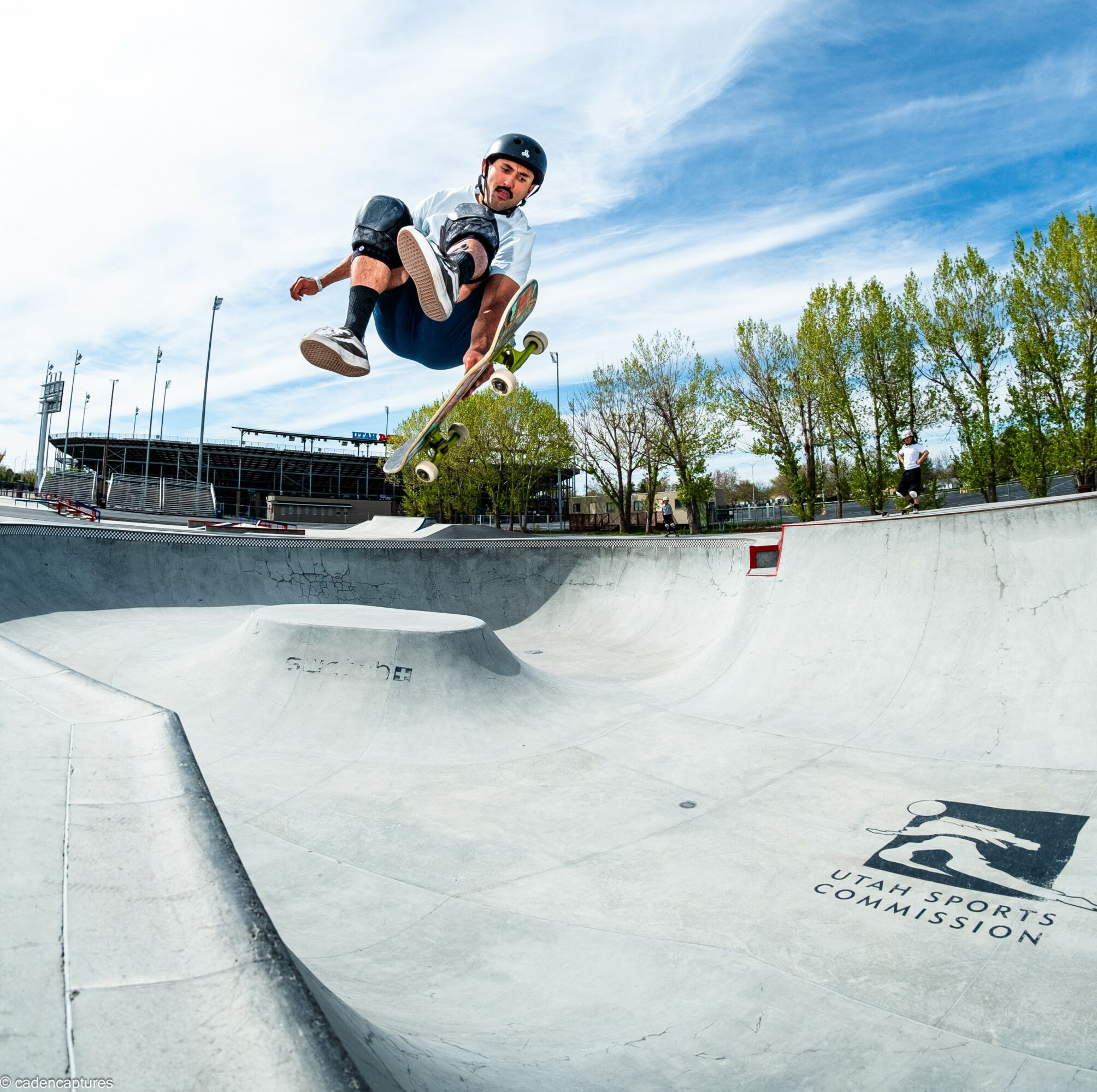 Skateboarder mid-air performing trick in skatepark with trees and blue sky in background.