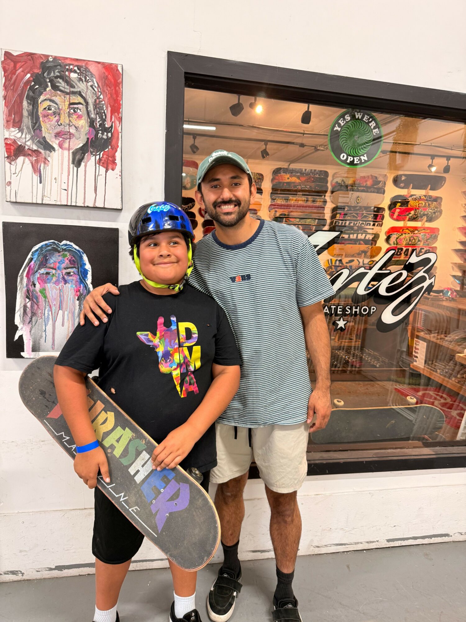 Two people, a young boy with a skateboard and an adult man, standing outside a shop with artwork on the wall.