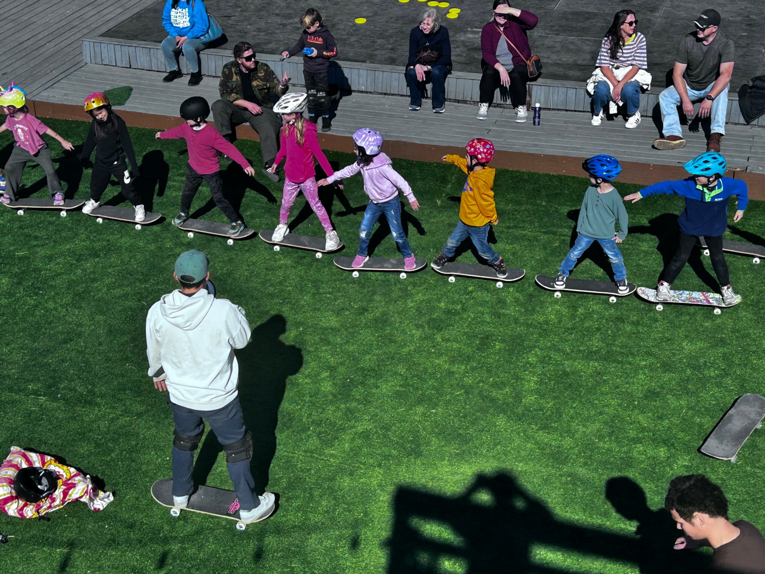 Children in colorful clothing ride skateboards in a line on a grassy area, with spectators sitting on benches nearby.