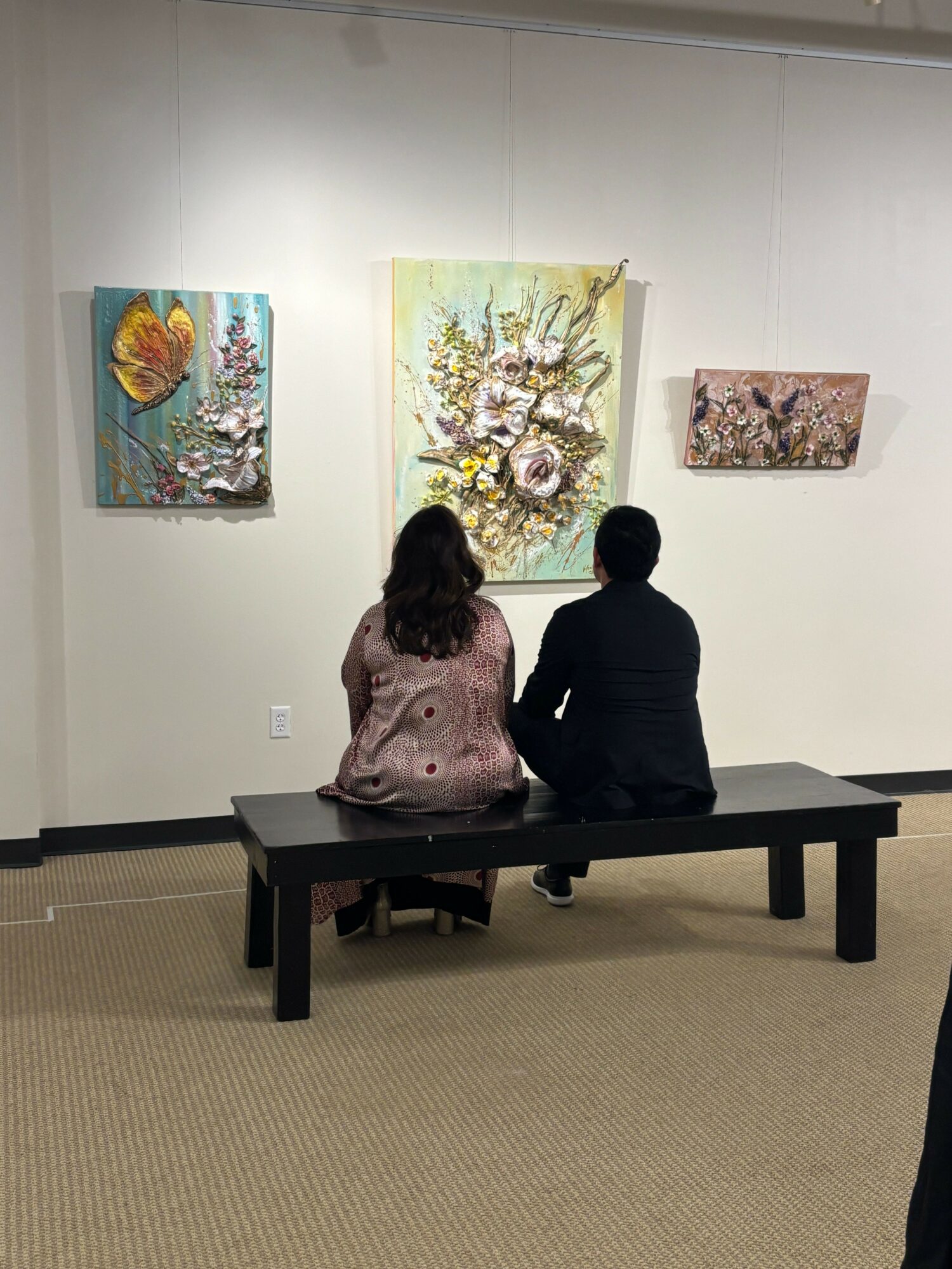 Two people sit on a black bench in an art gallery, viewing three paintings on a white wall.