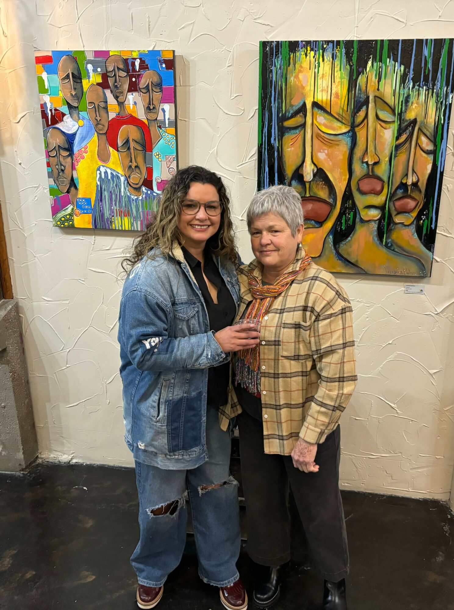 Two women stand together in front of colorful abstract paintings on a white wall, smiling at the camera.