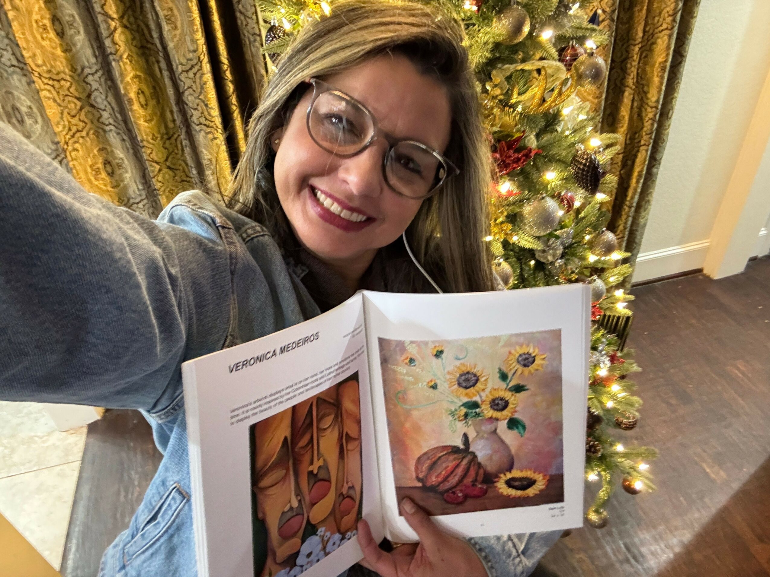 Woman with glasses smiling holding open book with colorful artwork, standing in front of decorated Christmas tree.
