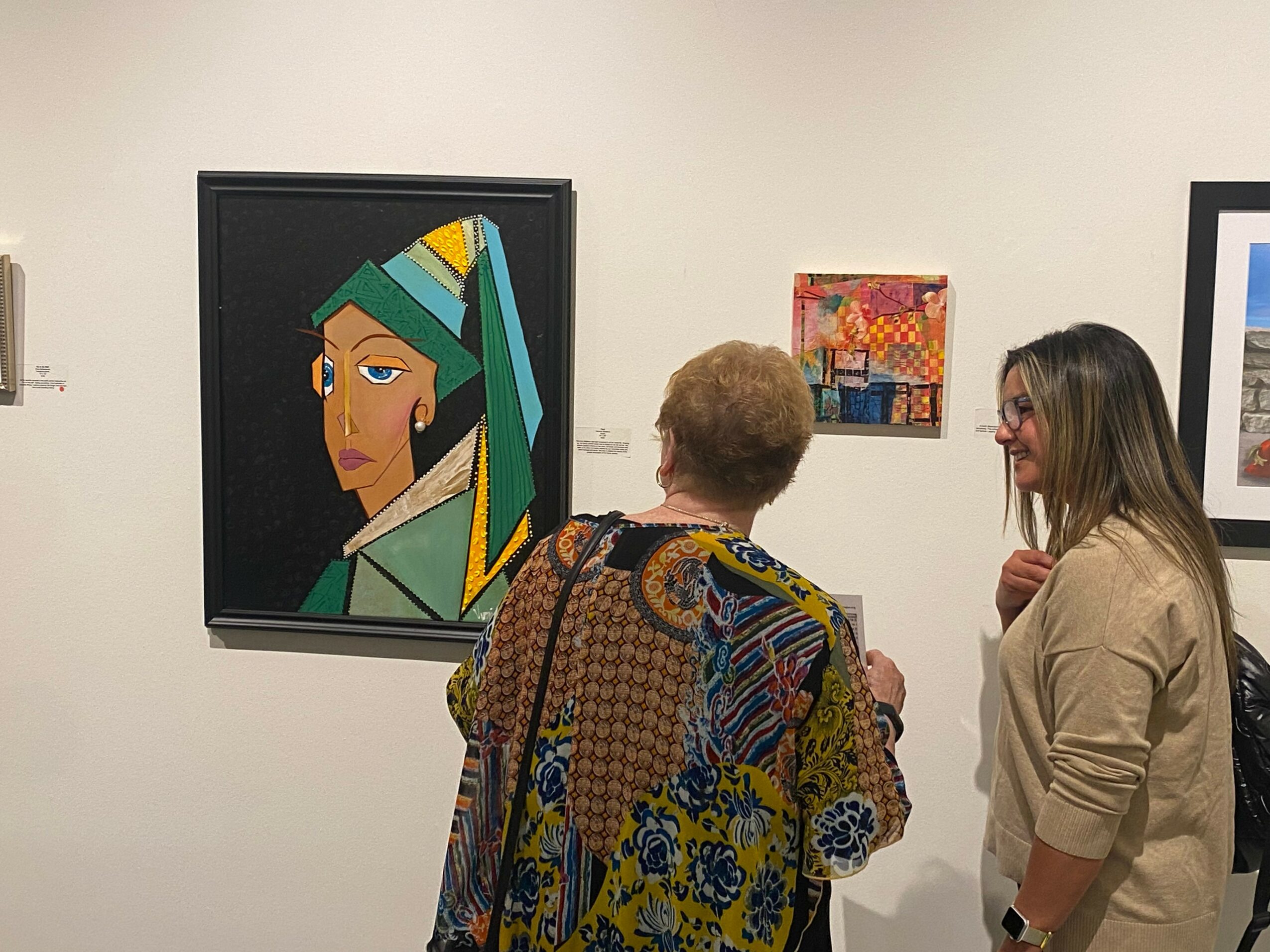 Two women looking at artwork in a gallery, one with short hair and patterned clothing, the other with long hair and beige jacket.