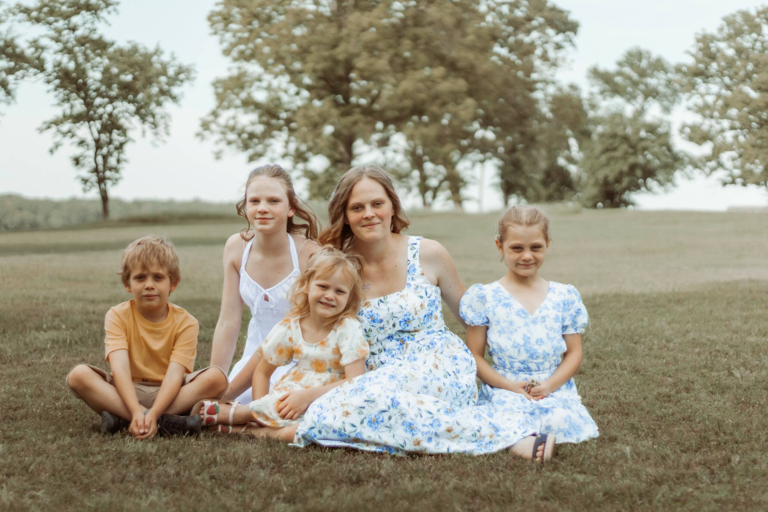 Group of five people, including children and an adult woman, sitting on grass outdoors with trees in background.