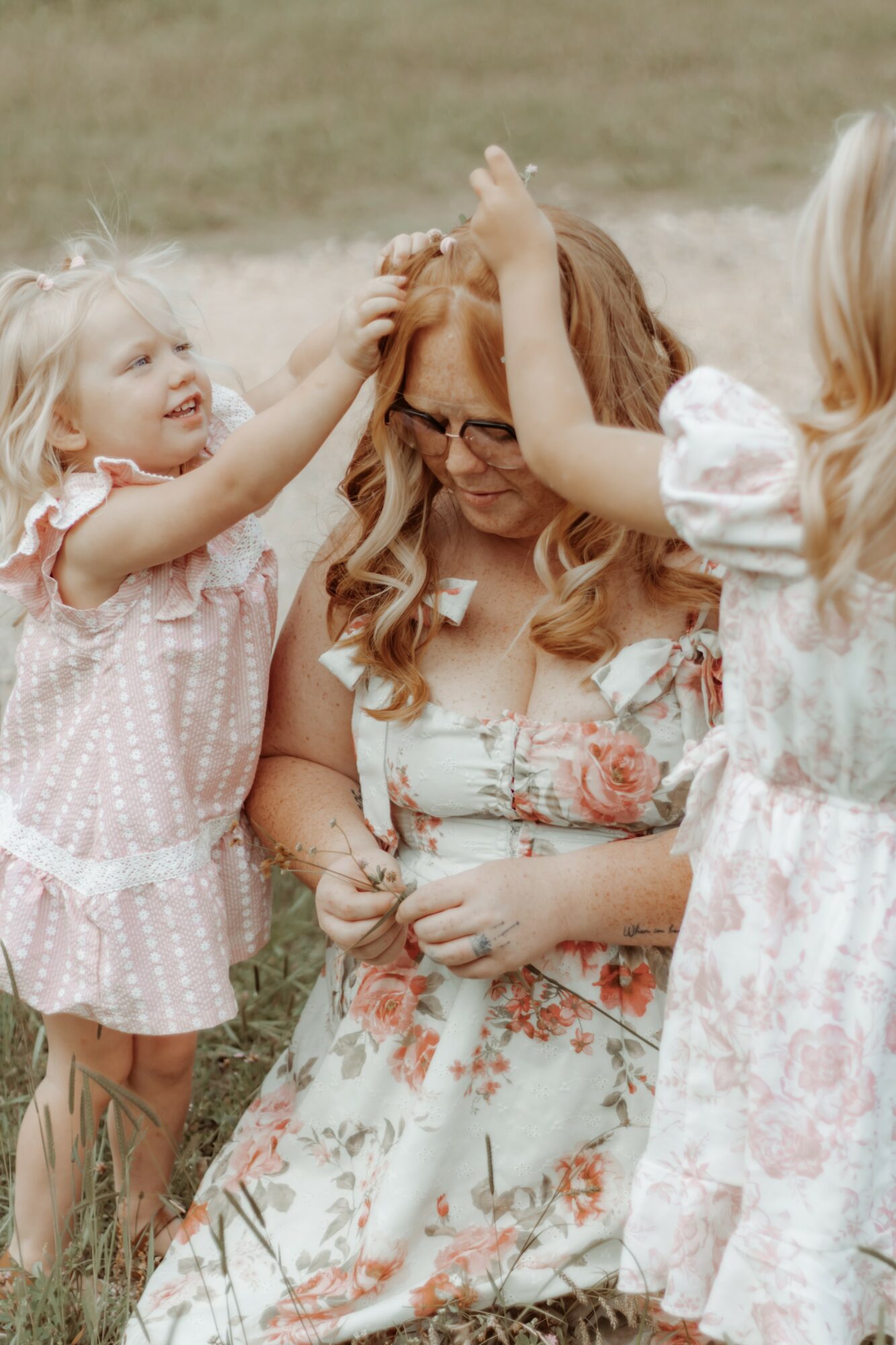 Two young girls playfully touch a woman’s head outdoors, all smiling and dressed in light-colored, floral-patterned clothing.
