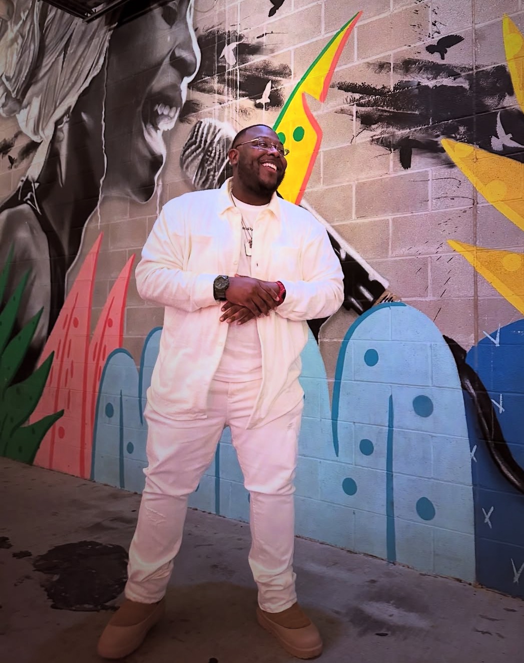 Person smiling and standing in front of colorful graffiti wall, wearing white outfit and beige shoes.