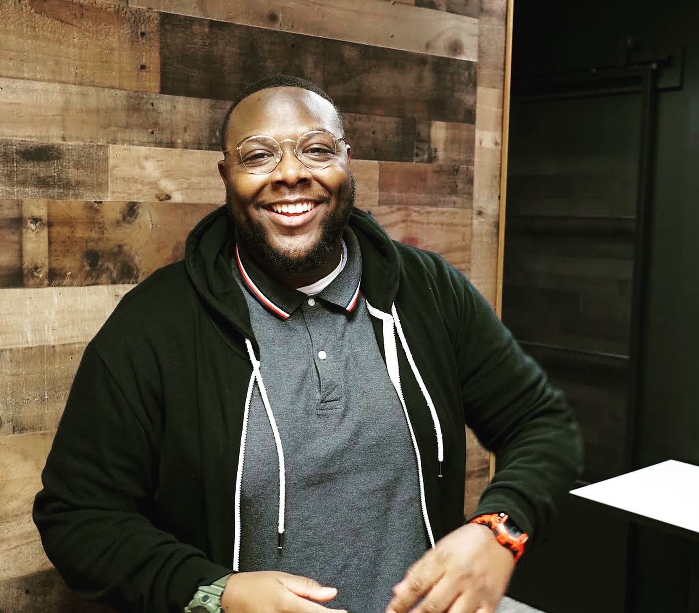 Smiling man with glasses, beard, wearing a hoodie and polo shirt, standing indoors with wooden wall background.